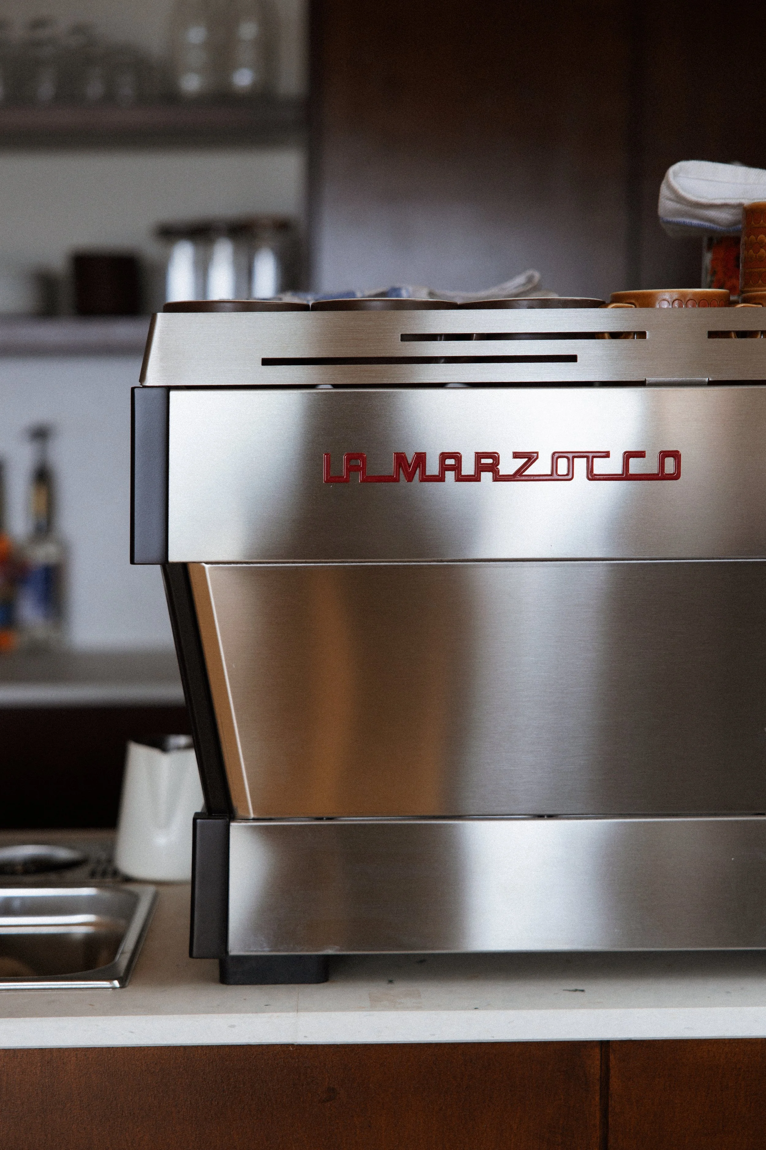 Close-up of a stainless steel La Marzocco espresso machine on a kitchen countertop, with cups and kitchen items in the background.