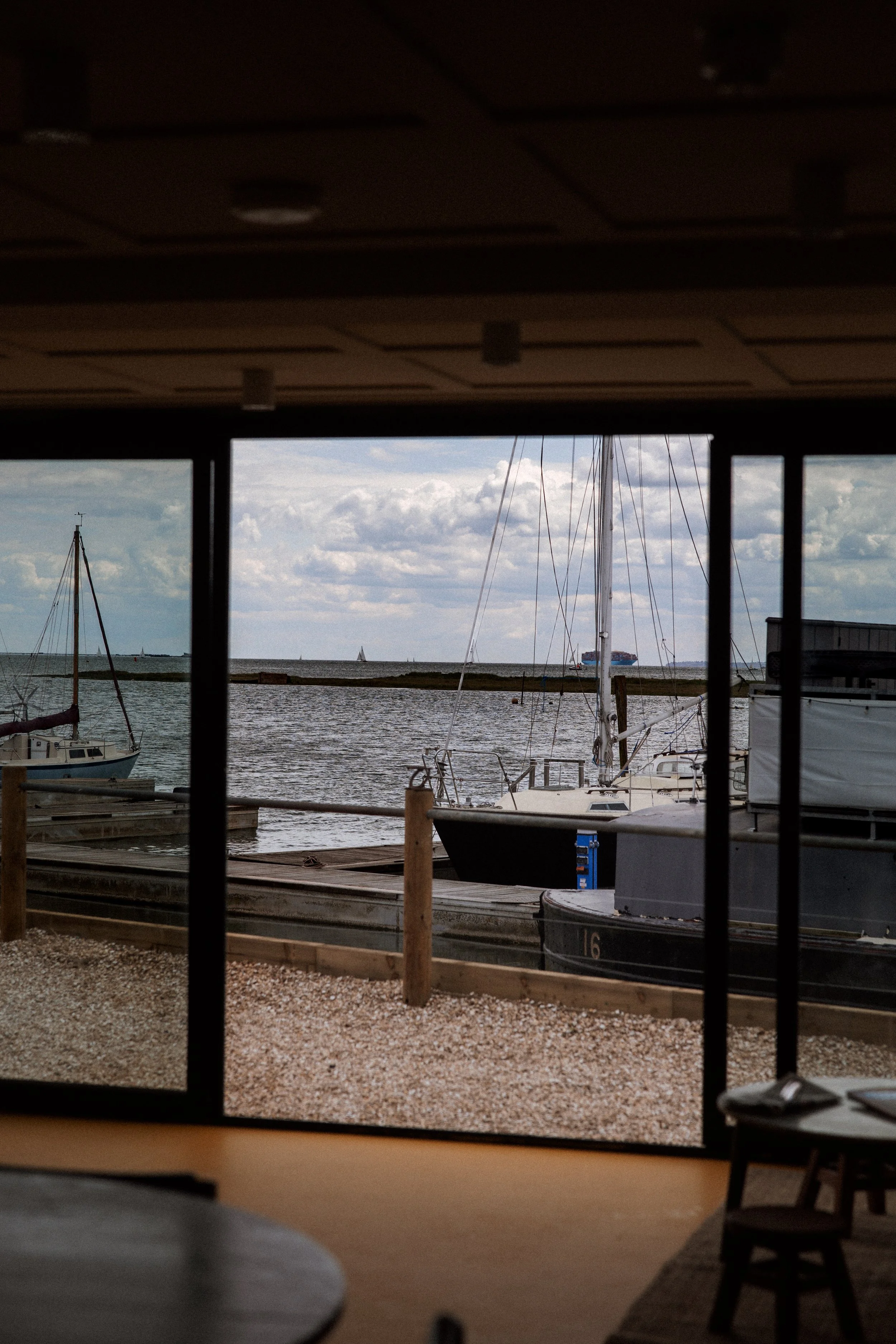 View of sailboats docked at a marina seen through large windows from inside a building with a ceiling and dark window frames, overlooking a body of water with the sky and clouds above.