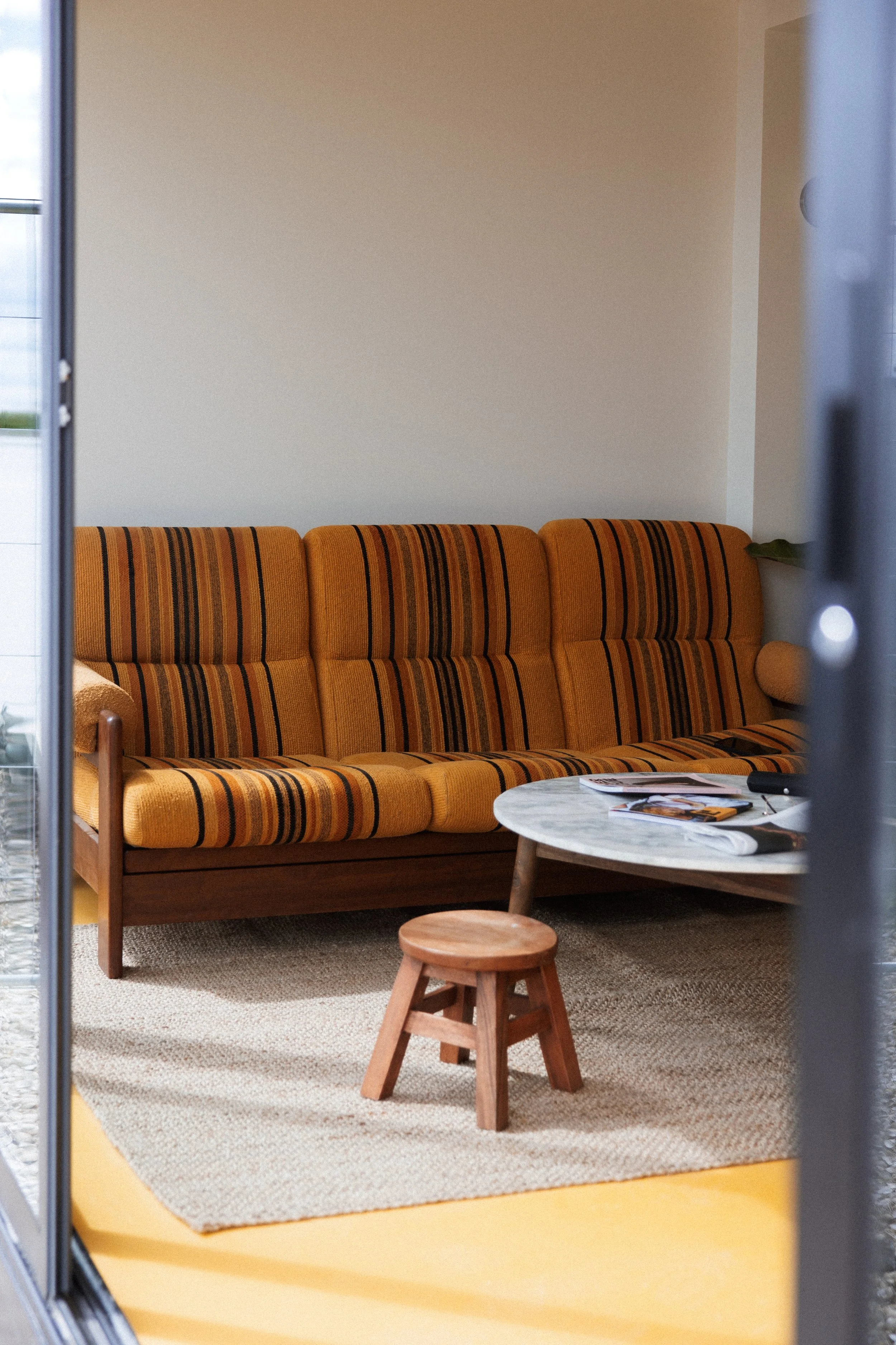 A vintage orange striped sofa with a wooden frame, a small wooden stool, and a round marble-top coffee table with magazines on it in a cozy living room. The room has a beige carpet and white walls, with a plant visible in the back corner.