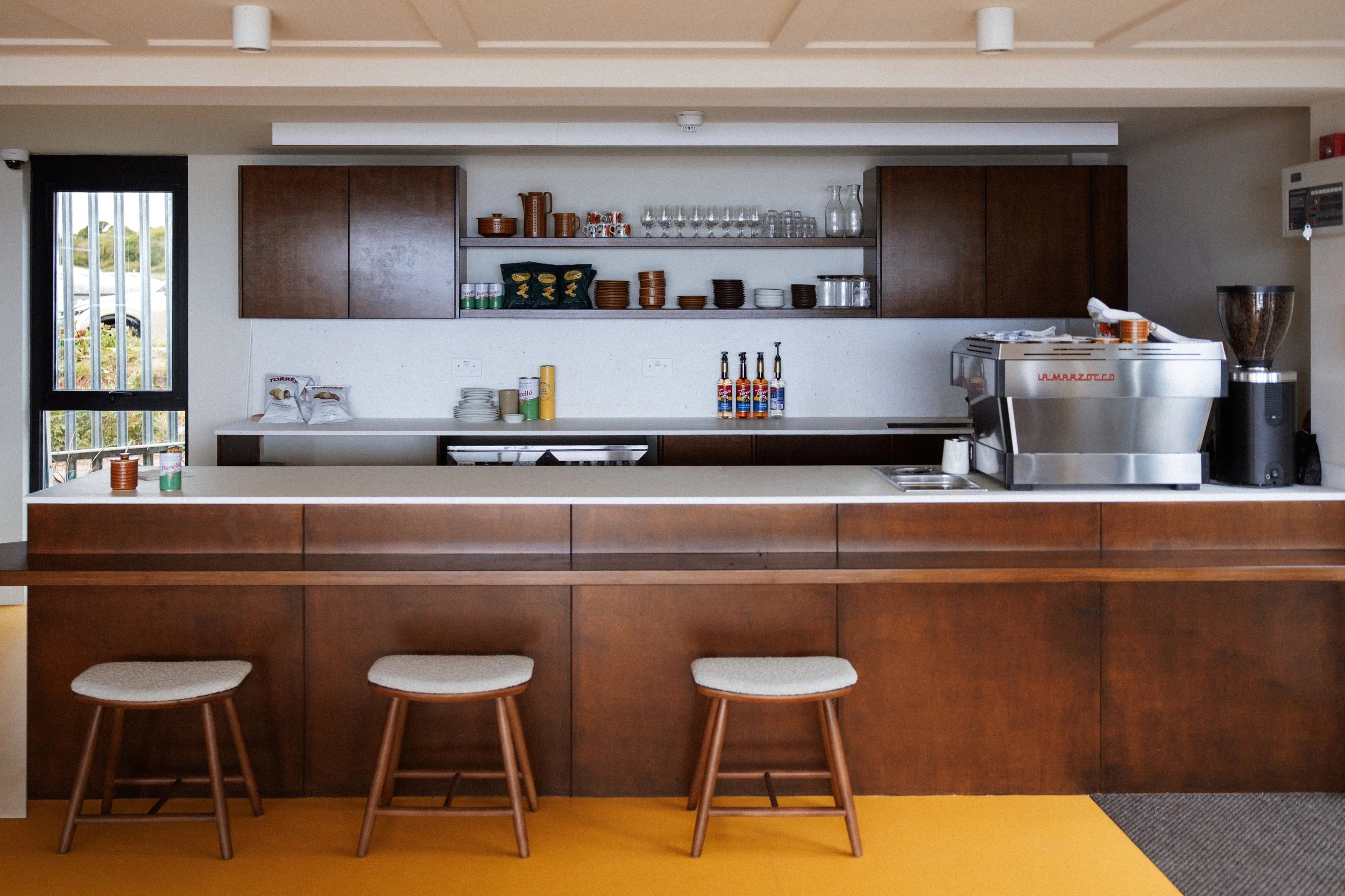 Modern kitchen with wooden cabinets, open shelf with glassware, coffee machine, stools, and a yellow floor.