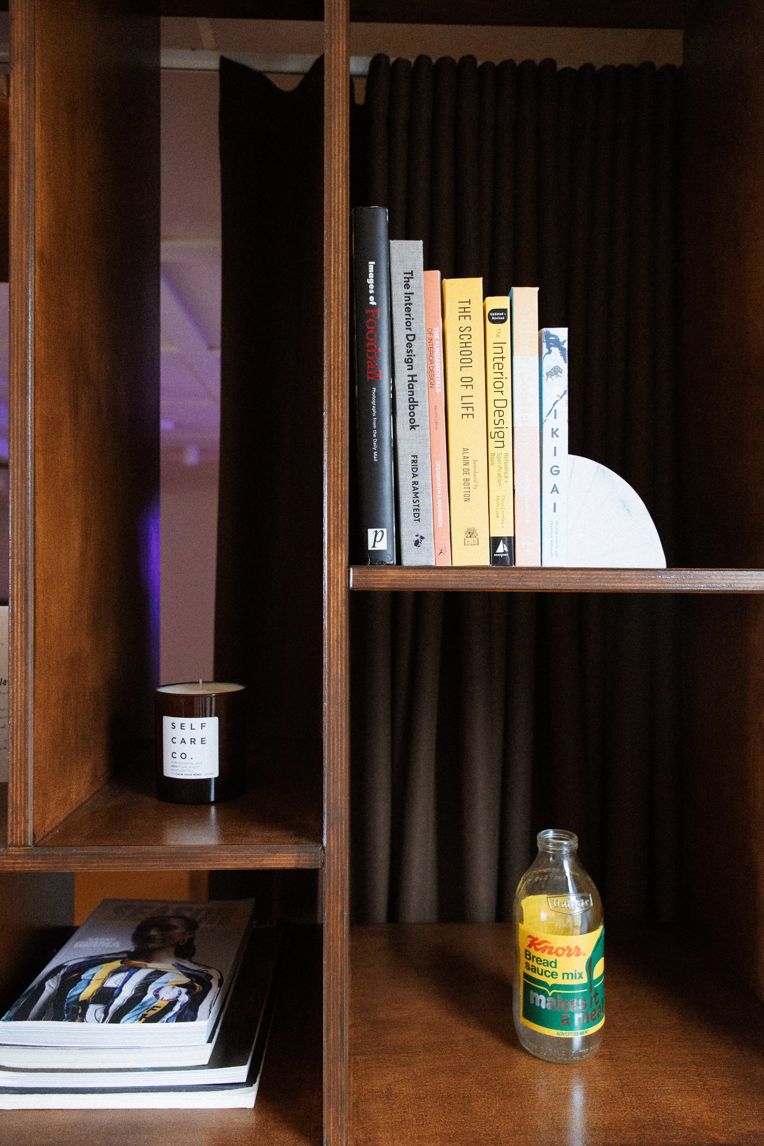 A wooden shelf with books on design and life, a black candle labeled 'Self Care Co.', and a bottle of Knorr Bread Sauce Mix. The background has dark curtains.