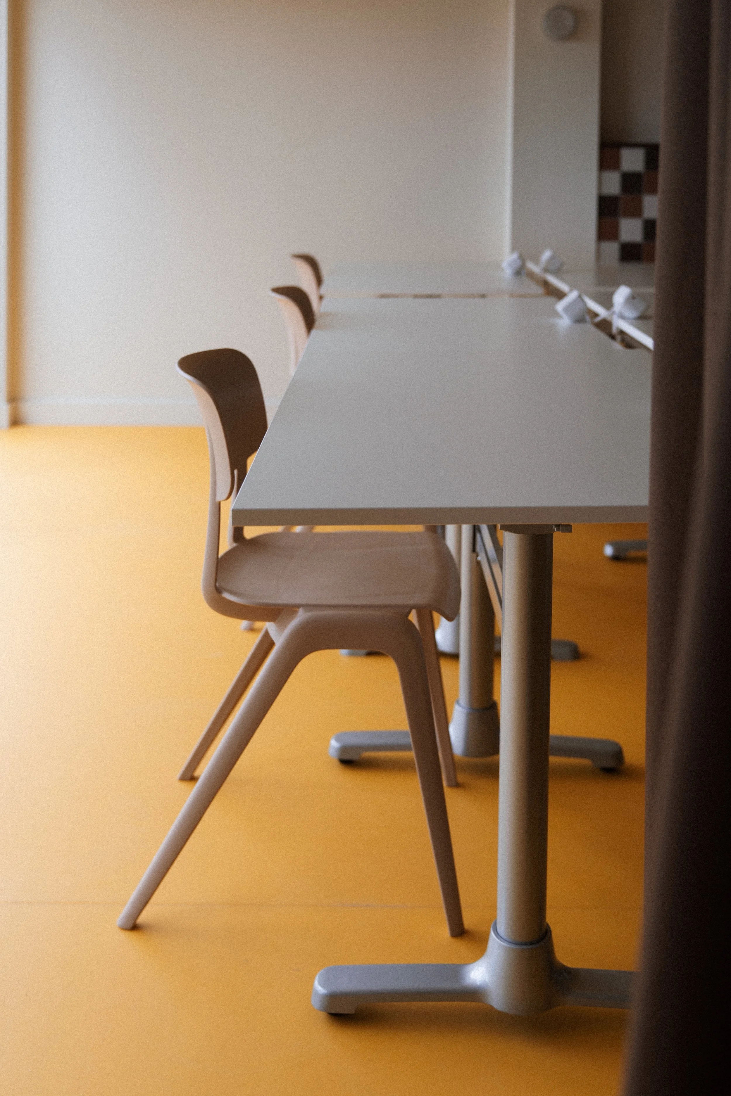 Empty chairs lined up along a white table in a room with a yellow floor.