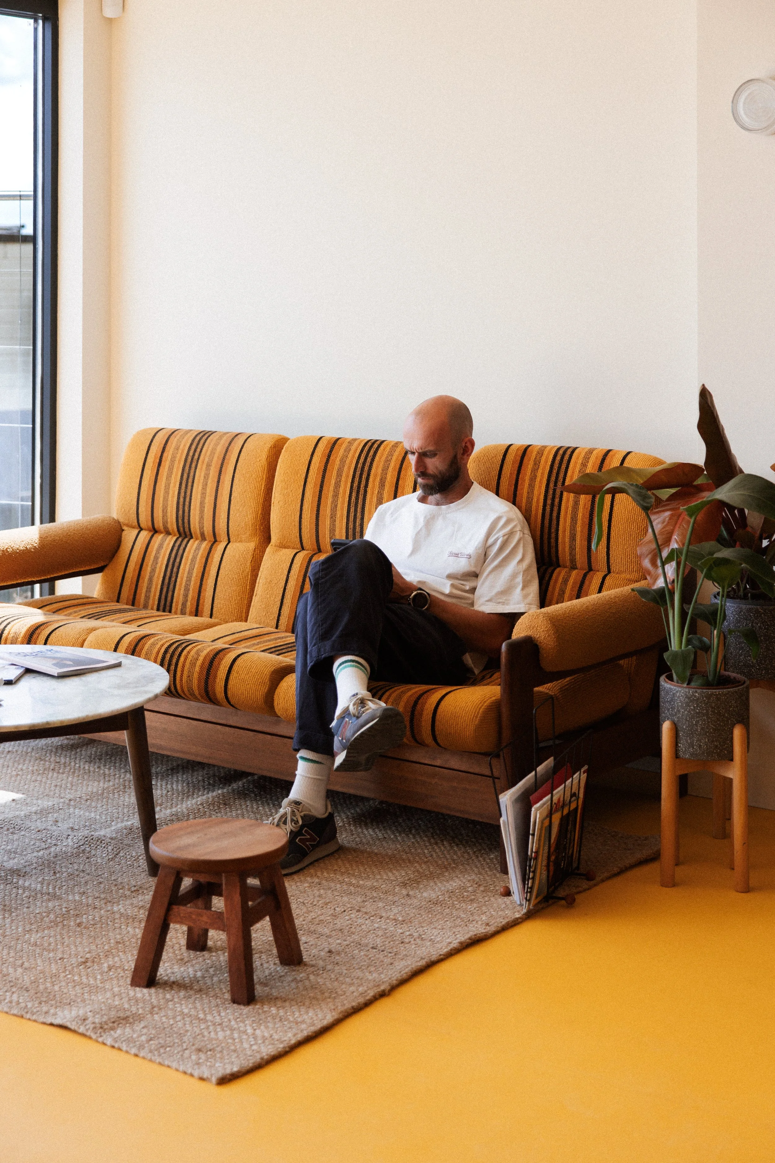A man with a shaved head and beard sitting on a yellow striped sofa, looking at his phone, in a room with a large window, a potted plant, and a small wooden stool.