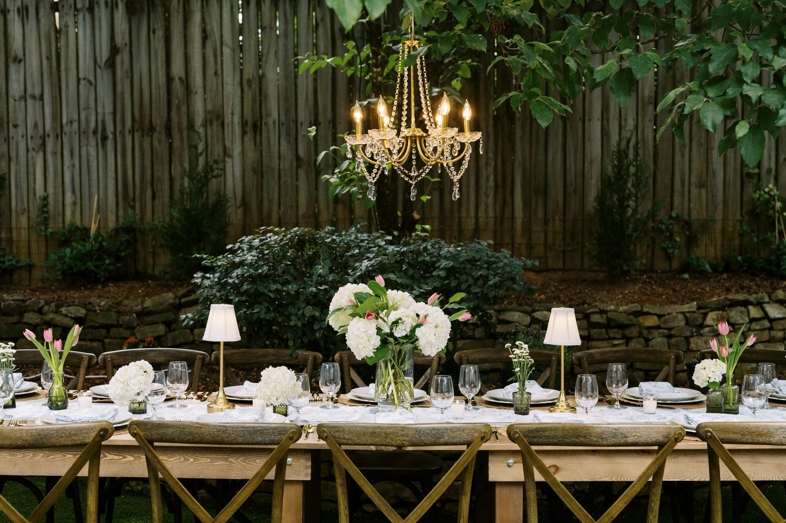 Elegant outdoor dining setup with a long table decorated with white floral centerpieces, surrounded by wooden chairs, with a chandelier hanging above, and surrounded by greenery and a wooden fence.