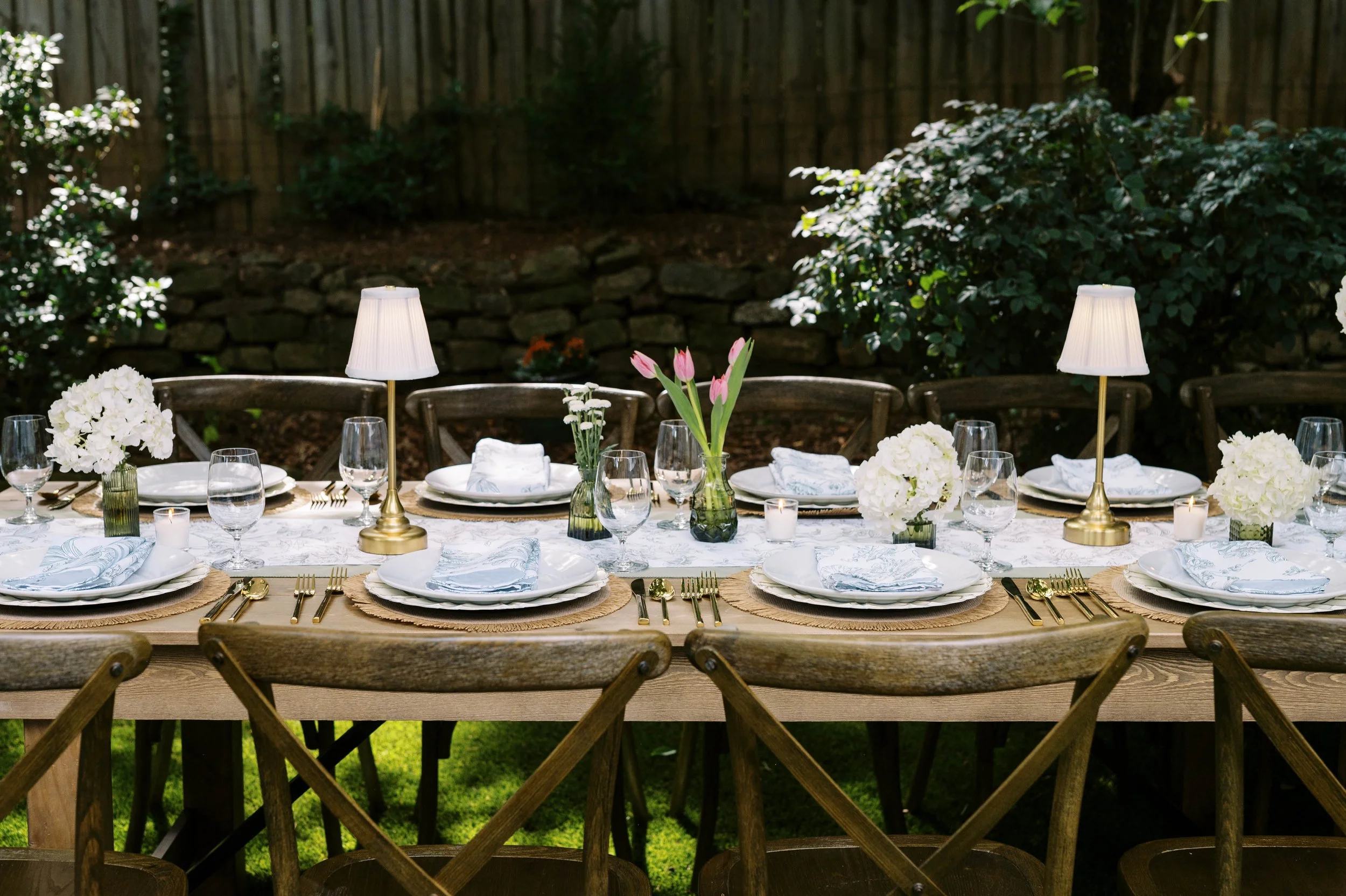 A decorated outdoor dining table set with white flowers, pink tulips, candles, glassware, and gold flatware, with wooden chairs around it, in a garden setting with greenery. Rental items available in Decatur and Atlanta, GA.