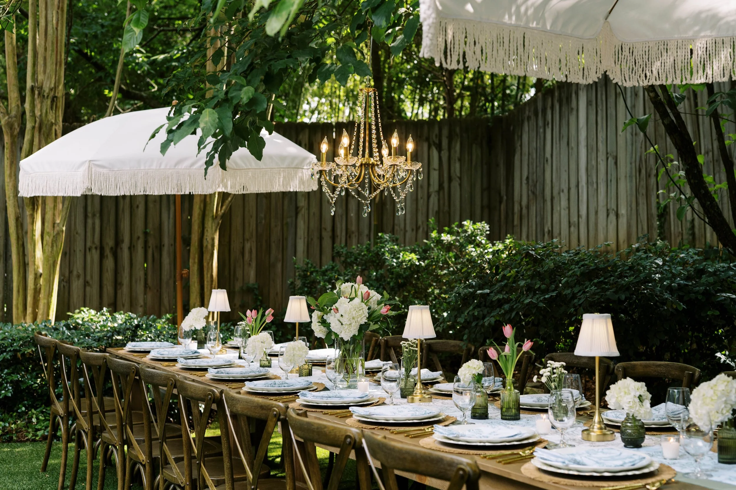 An outdoor dining setup with a long table decorated with white flowers, gold accents, surrounded by wooden chairs, with warm light lamps and umbrellas providing shade. Boutique event rentals in Decatur, GA.