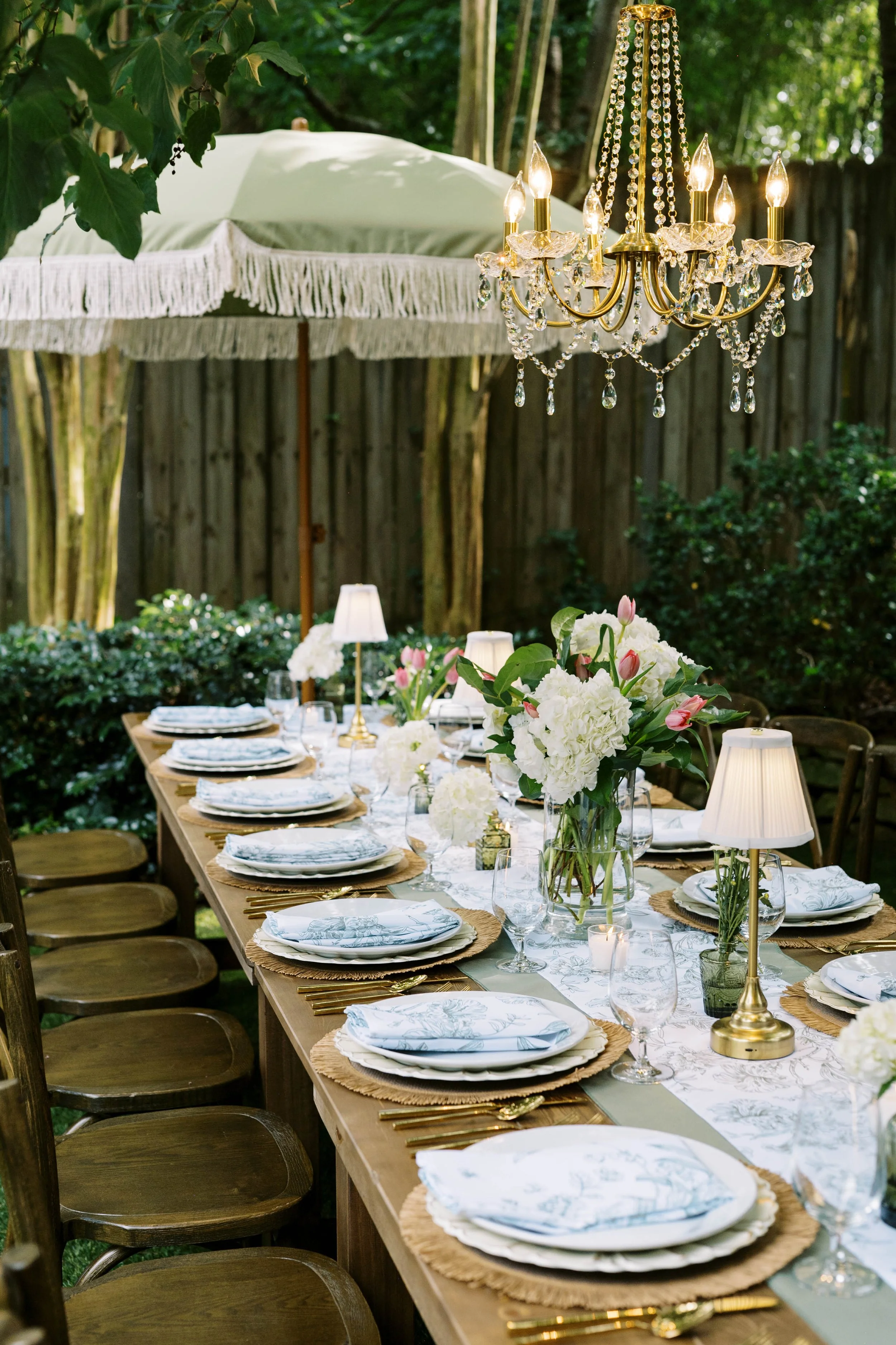 An outdoor dining table decorated with white floral tablecloths, surrounded by wooden chairs, and set with plates, glassware, gold flatware, and white napkins. There are floral centerpieces with white and pink flowers, table lamps, and a crystal chan