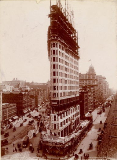 A tall building under construction in a busy city street with cars and pedestrians, featuring scaffolding at the top.