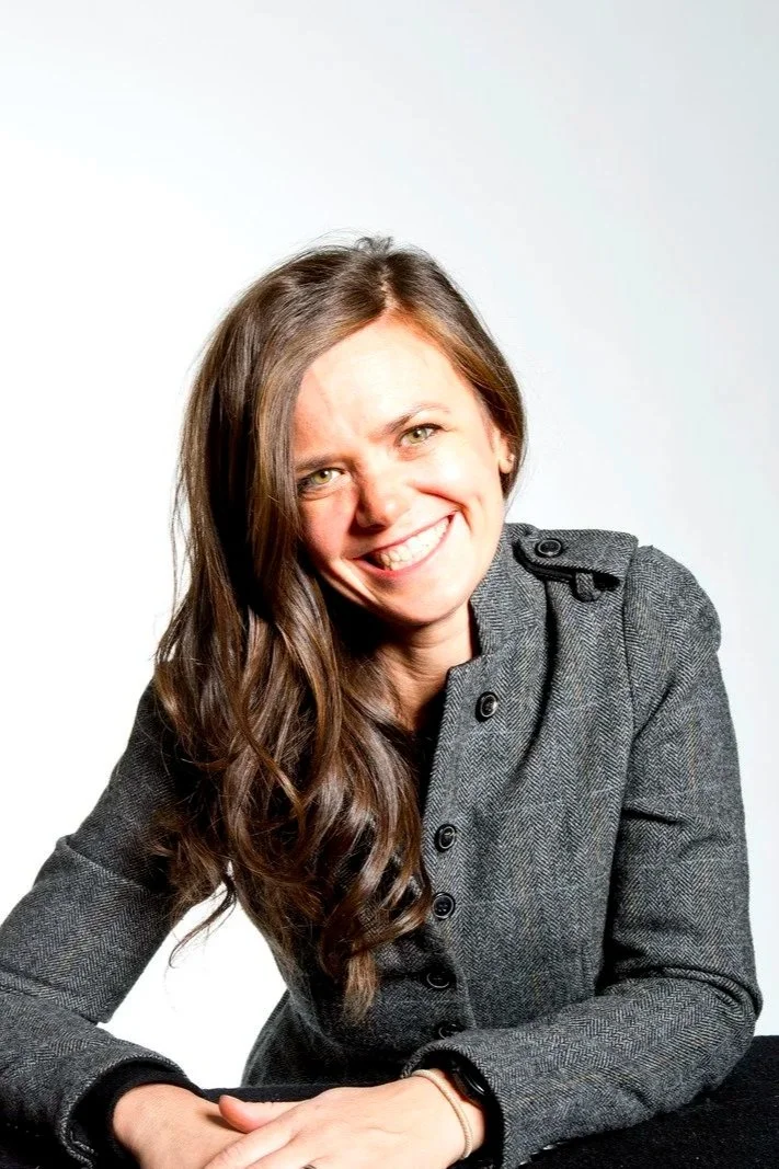 A young woman with long brown hair smiling, wearing a gray blazer, sitting at a table with a white background.