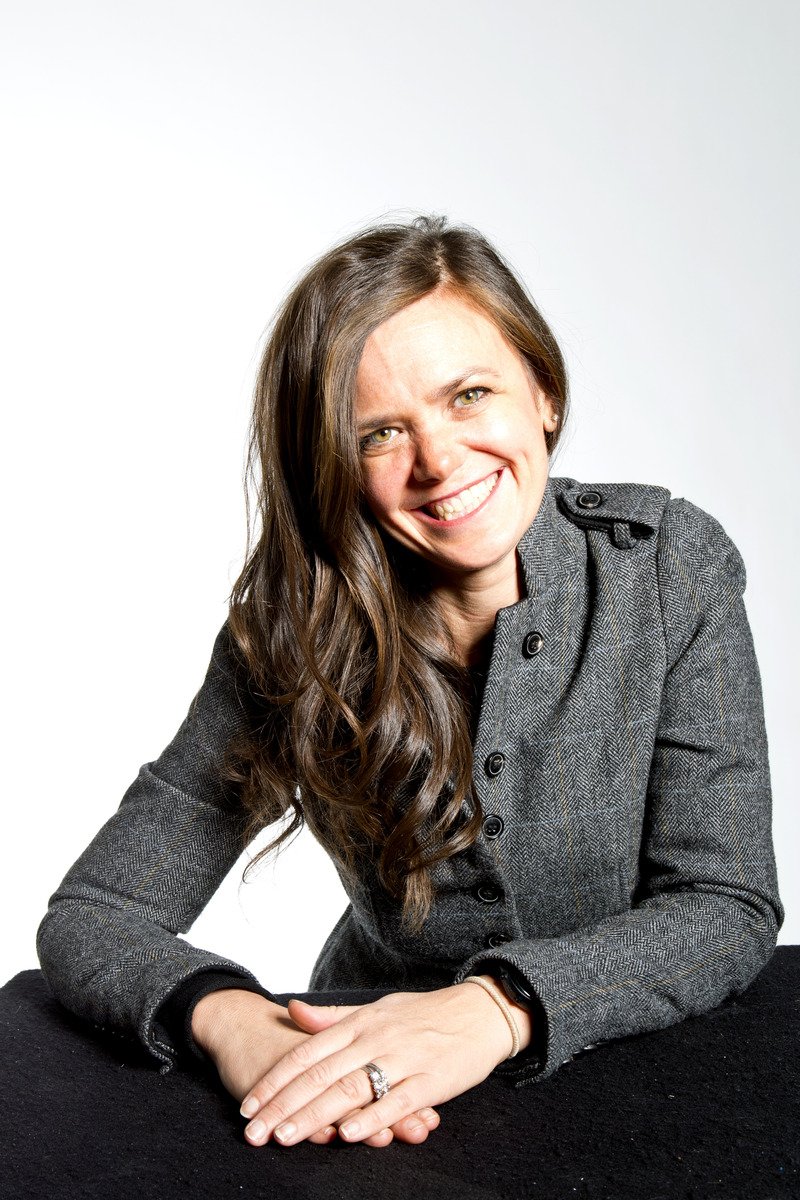 A woman with long brown hair smiling while sitting at a table, wearing a gray blazer and a ring on her left hand against a plain white background.