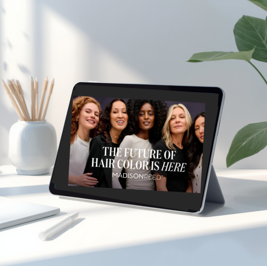 Digital tablet displaying an advertisement for a hair color brand featuring a diverse group of women with different hair colors and styles, on a white desk next to a plant and stationery.