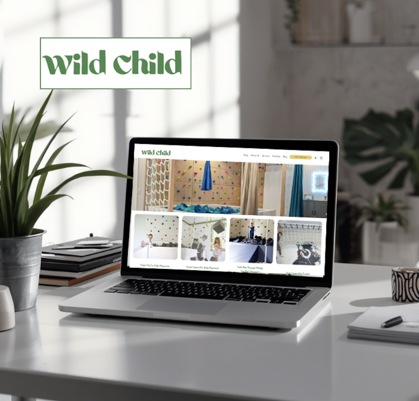 A laptop on a white desk displaying an online store called 'Wild Child' that features indoor climbing gyms and play areas for kids. The desk also has a potted plant, a stack of notebooks, and a pen. The background shows a bright indoor space with plants and a window.