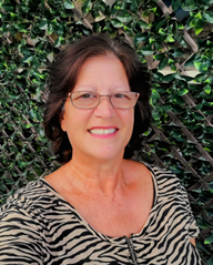 A woman with short dark hair and glasses smiling in front of a leafy green background, wearing a zebra print top.