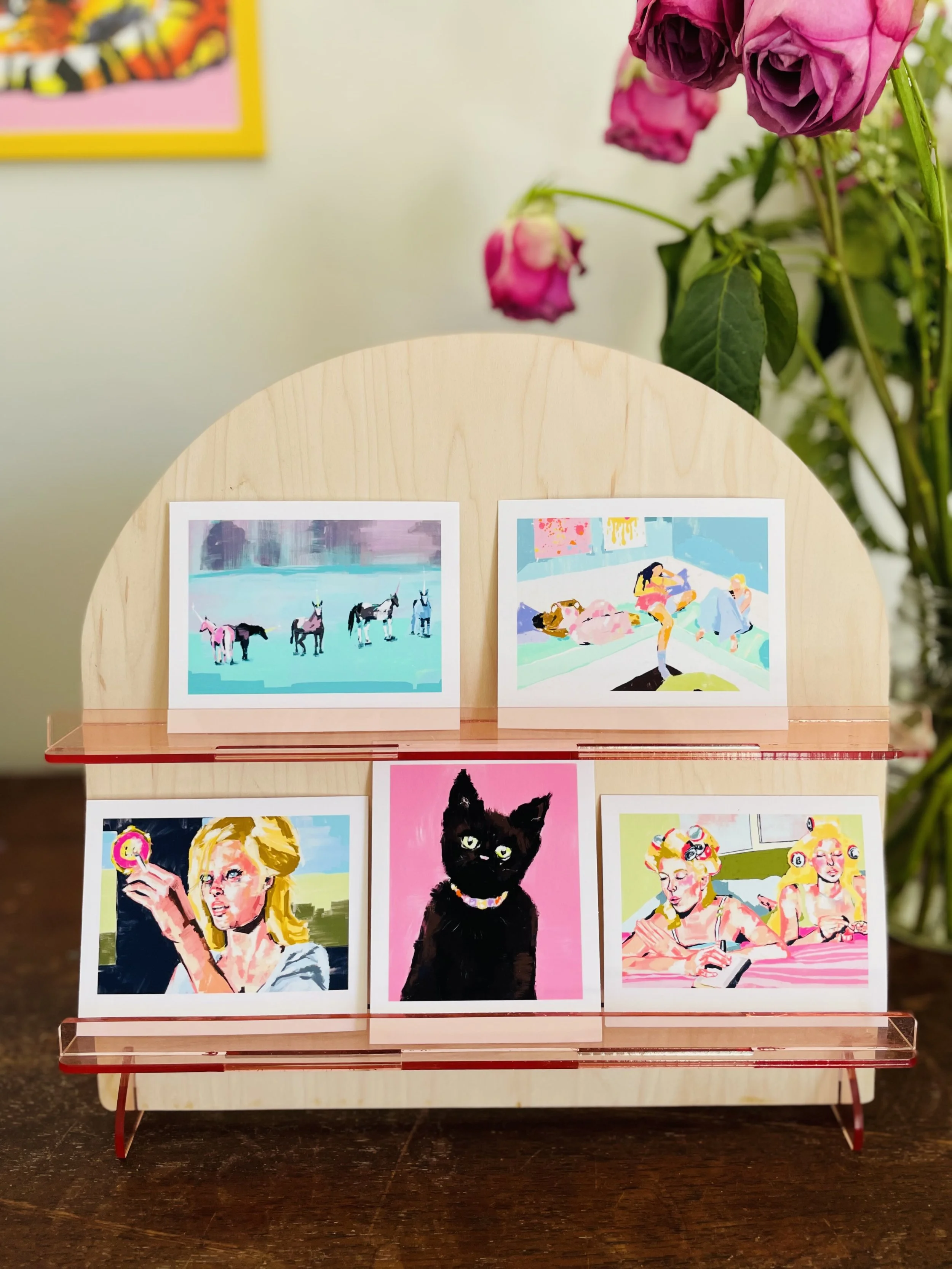 Colorful artwork displayed on a small wooden stand featuring four paintings, with pink roses visible in the background.