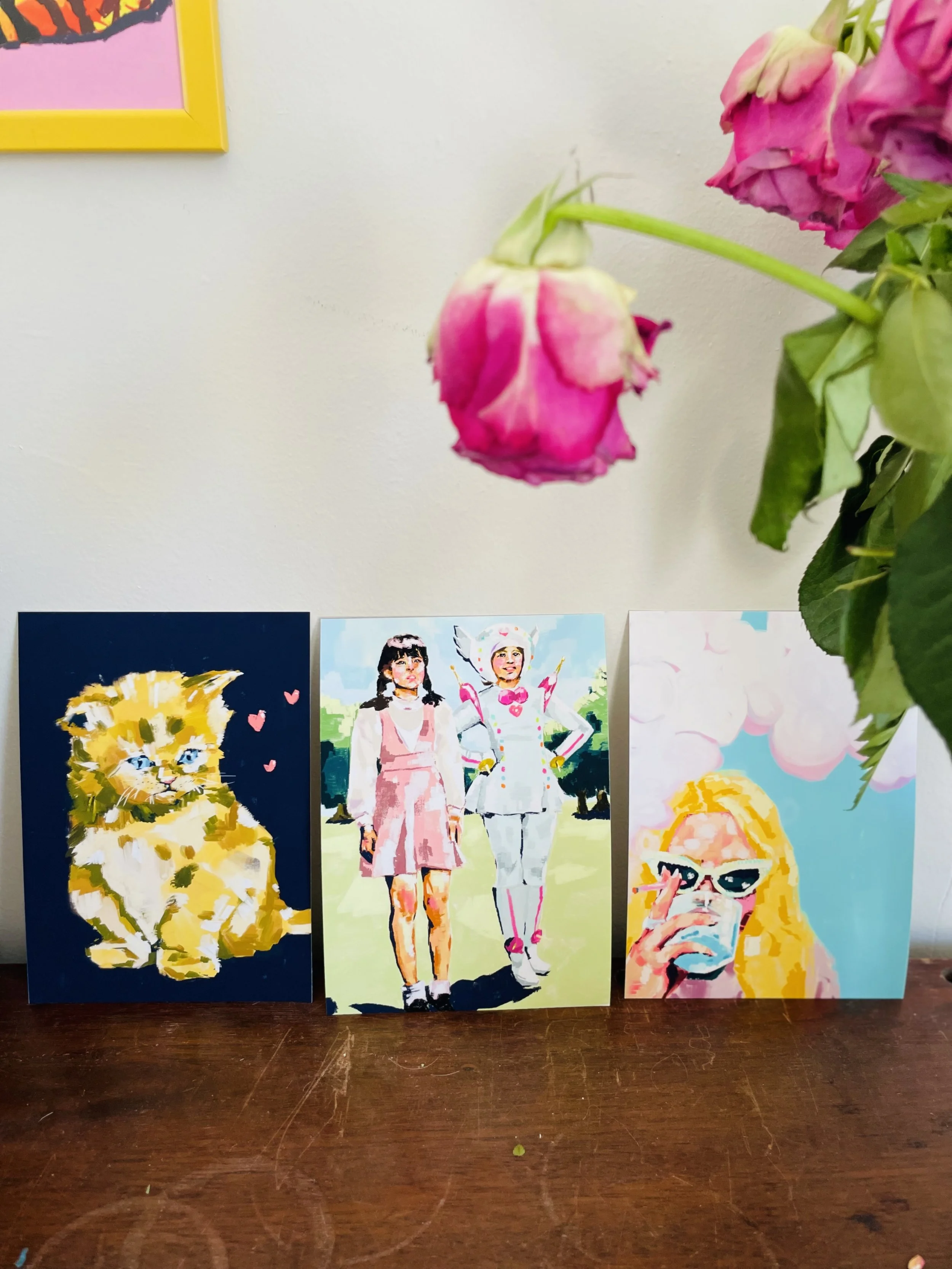 Three colorful posters featuring stylized portraits of children, placed on a wooden surface. One poster shows a yellow kitten with blue eyes, the middle poster depicts two girls in dresses, and the third features a girl with blonde hair and sunglasse