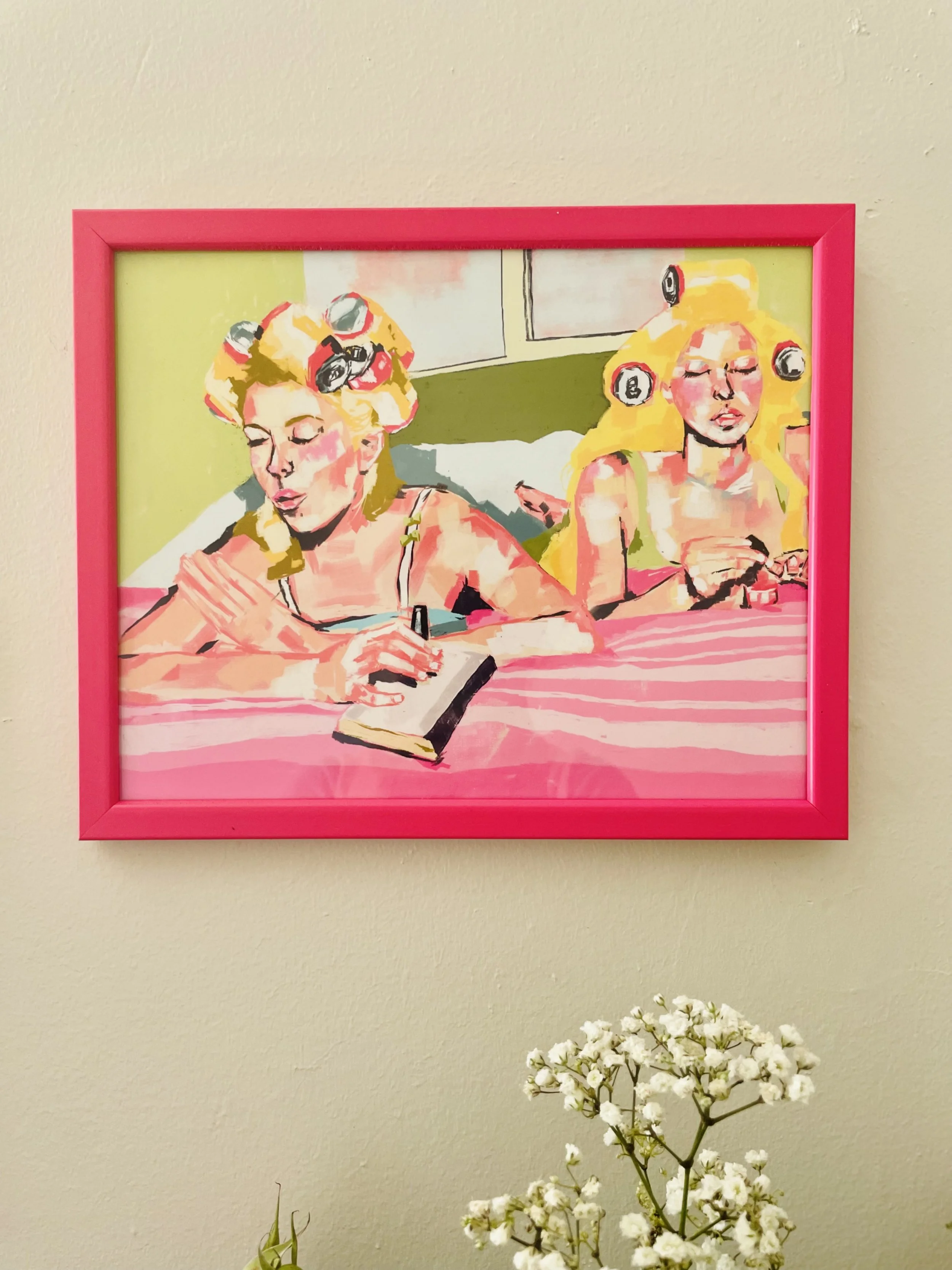 An art print framed in pink on a beige wall depicts two women with blonde hair, both with curlers in their hair. One woman is reading a book while the other is looking at her phone, with a window in the background. White flowers are in the foreground