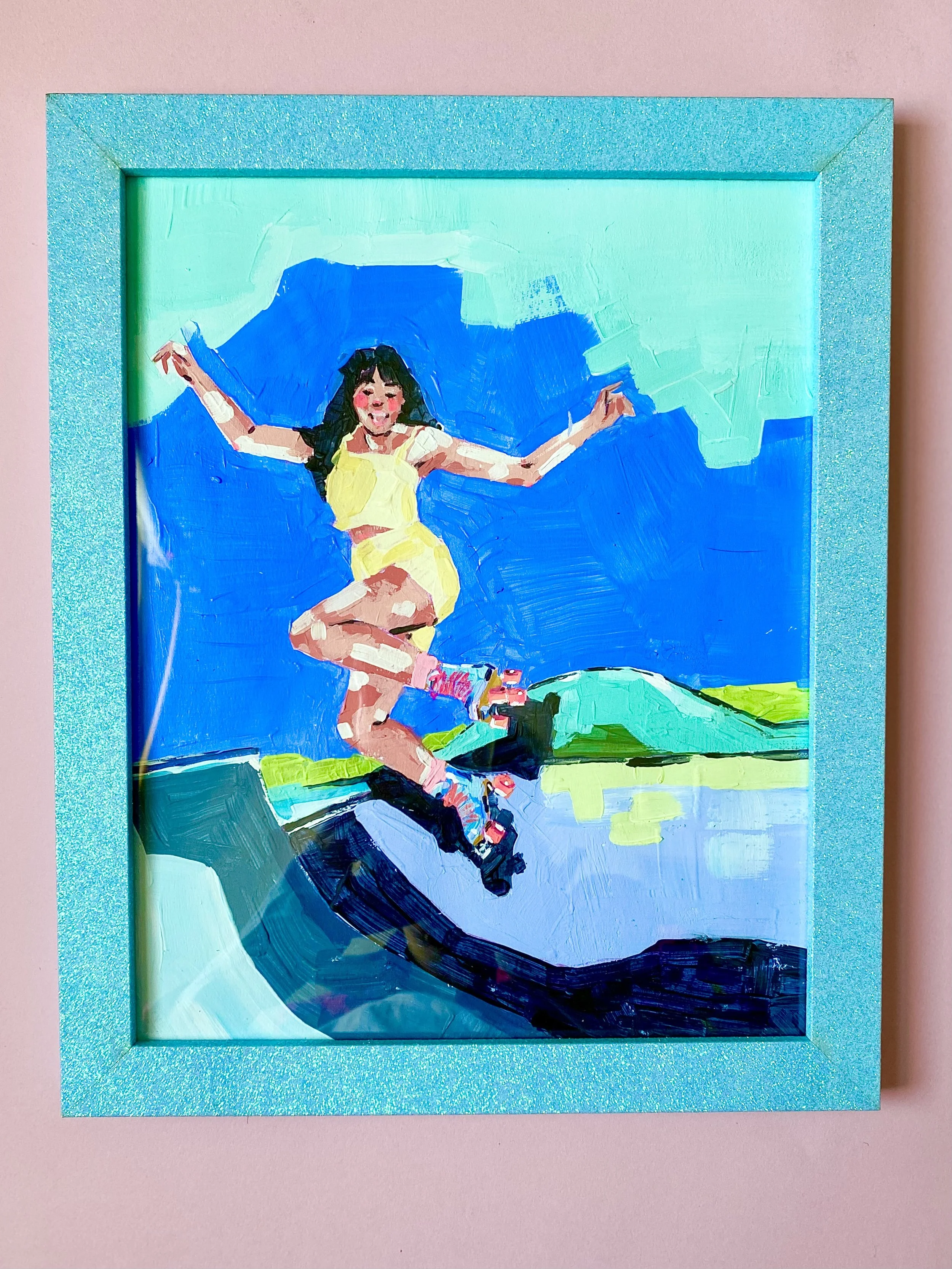 A colorful painting of a girl with dark hair in a yellow dress jumping with arms outstretched against a blue sky, framed in a sparkly blue frame, mounted on a pink background.