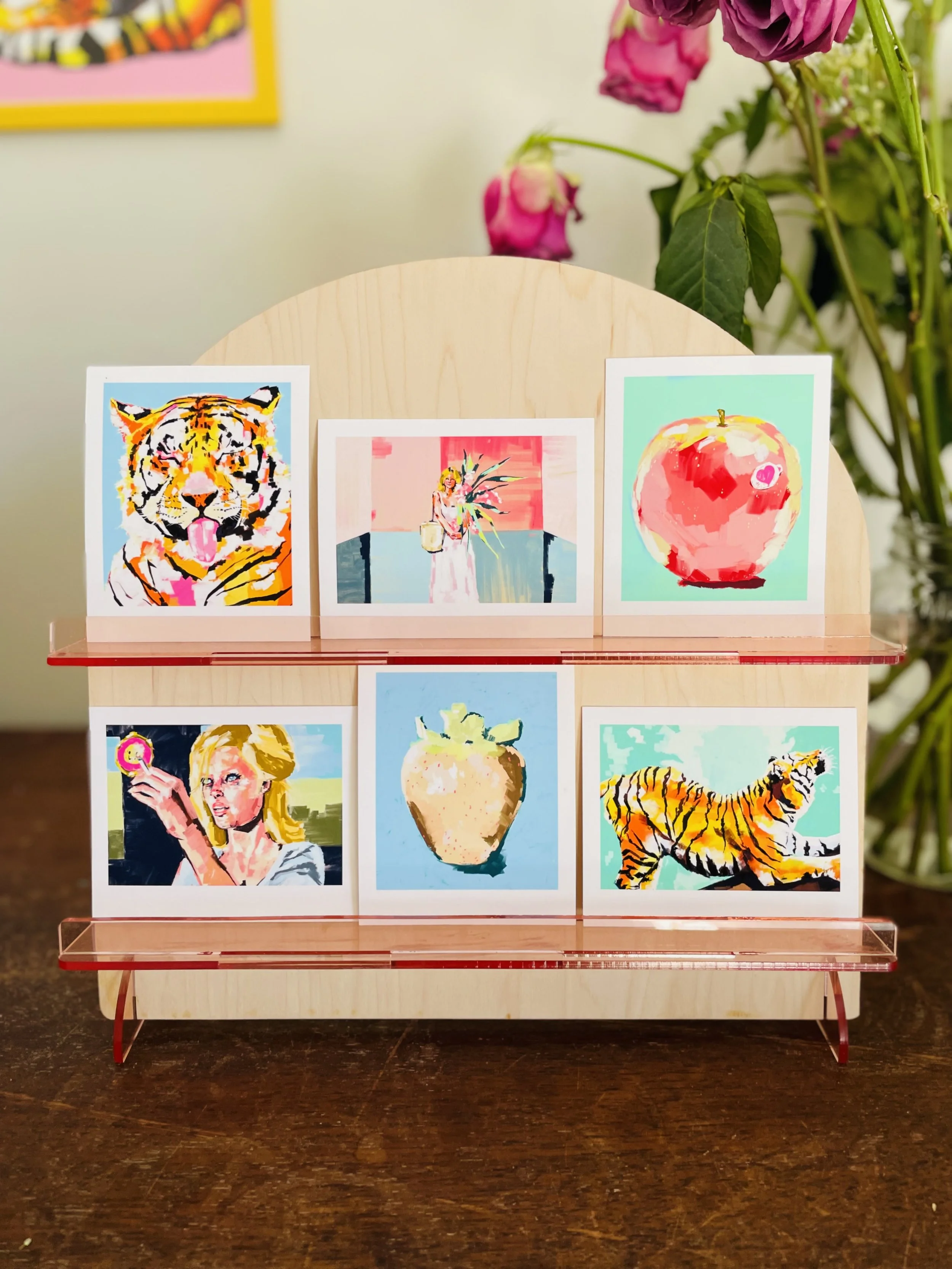 Six colorful art prints displayed on a wooden stand, featuring a tiger, a woman holding a plant, an apple with a heart, a woman with blonde hair, a vase with a plant, and a tiger lounging, with a plant with pink flowers in the background.