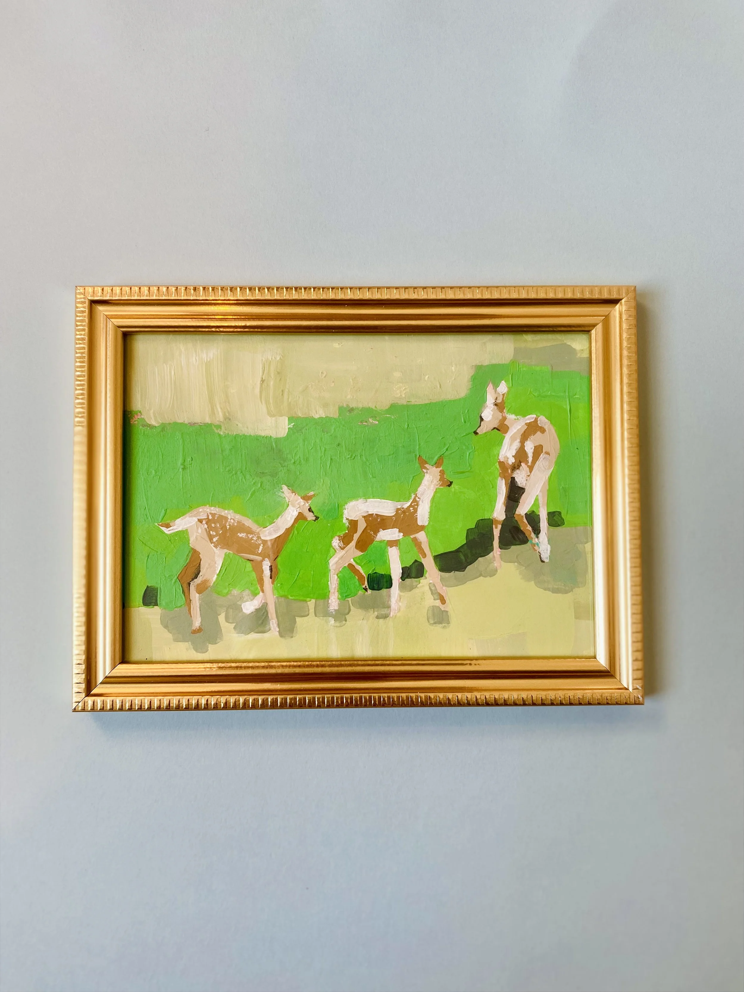 Painting of three deer, two fawns and an adult, standing on green grass with a yellow sky, in a wooden frame hung on a light blue wall.