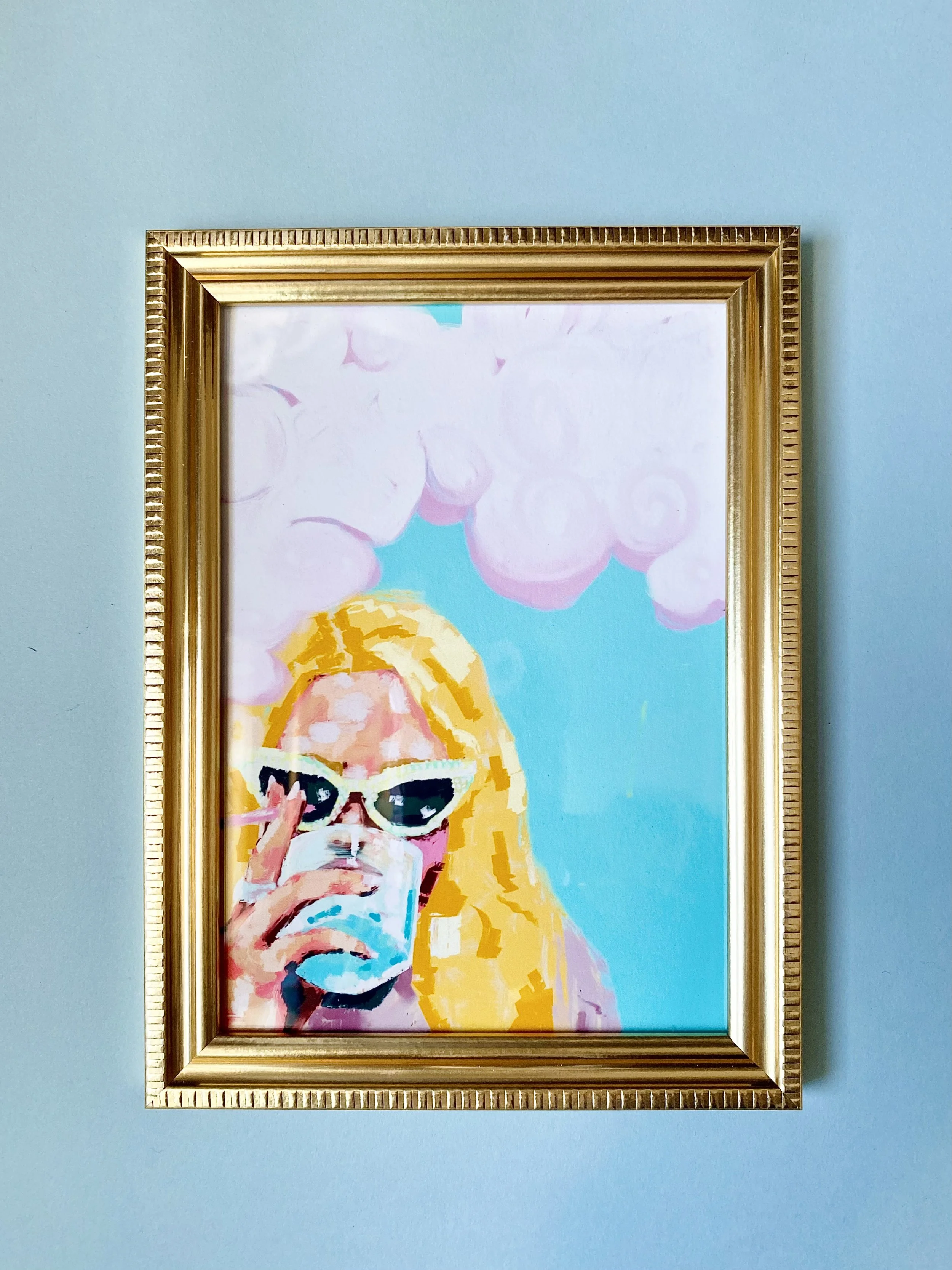 Painting of a woman with blonde hair wearing sunglasses and holding a glass, with pink clouds and a blue sky in the background, in a gold frame on a light blue wall.