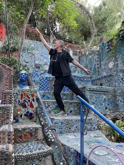 A man balancing on a mosaic-tiled staircase in an outdoor area with decorated mosaic walls, greenery, and sculptures. Location: The world famous Garden of Oz, Los Angeles, CA