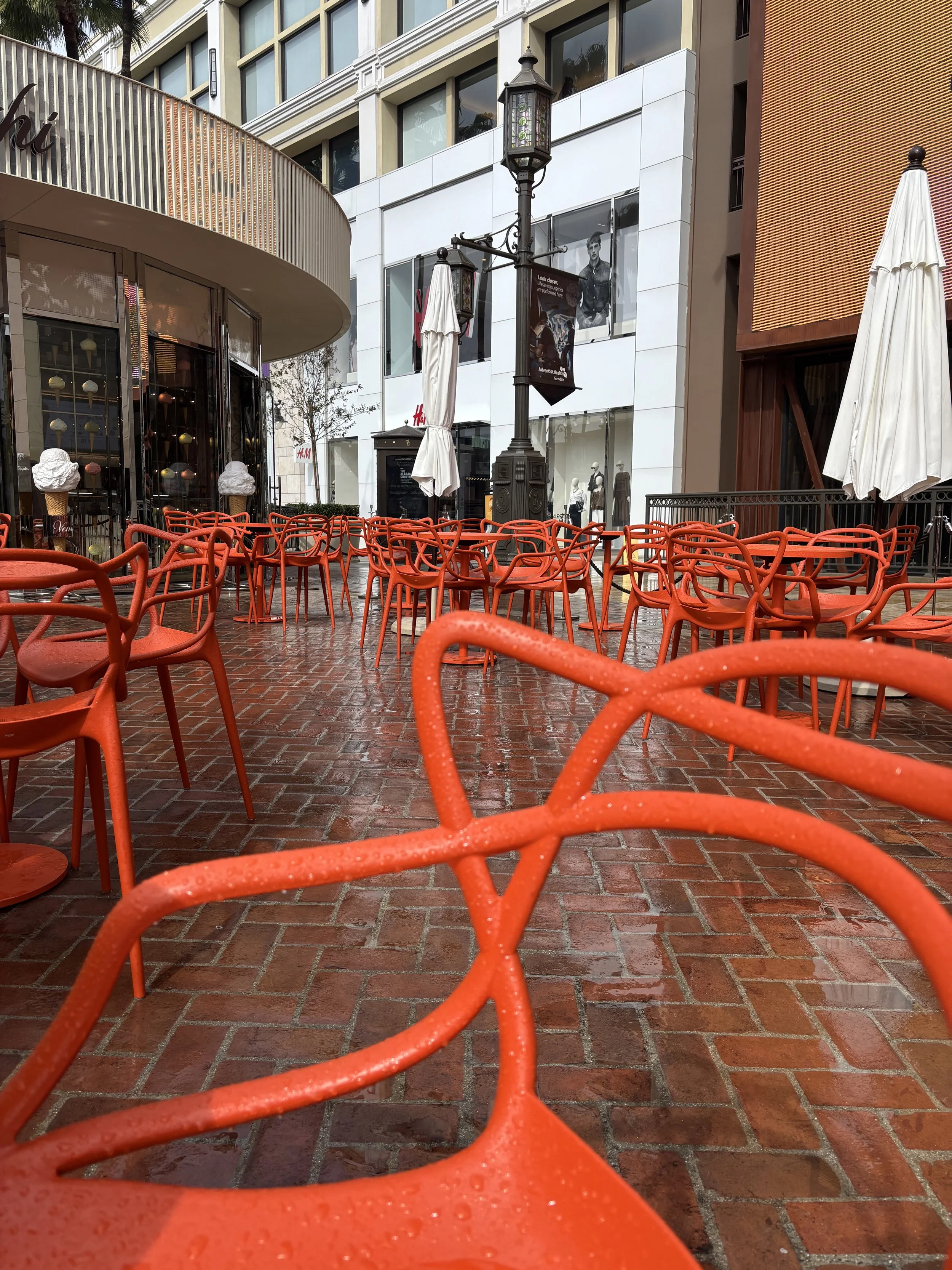 Empty outdoor patio with red chairs and tables, white umbrellas, nearby store with mannequins in windows, tall buildings, and a lamppost, possibly in a shopping district or mall.