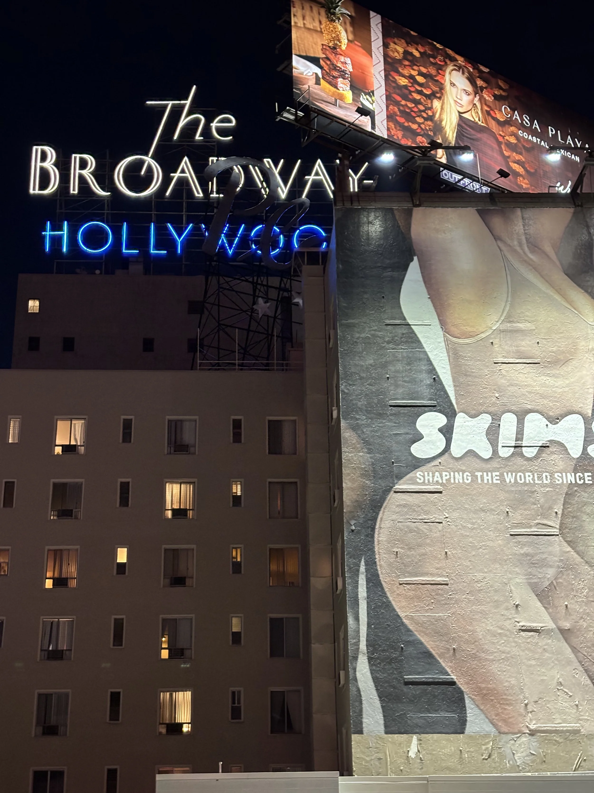 Nighttime photo of an illuminated sign for The Broadway-Hollywood, with a large billboard featuring a woman in front of a wall of orange flowers. The billboard advertises Casa Playa, a coastal Mexican restaurant, with images of grilled meat and pineapples.