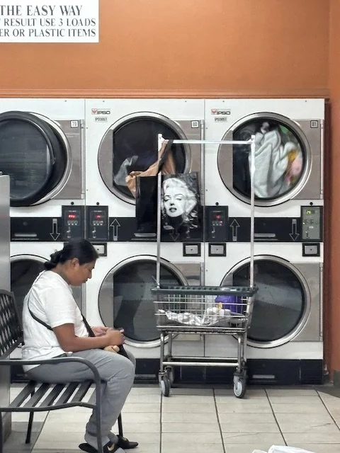 A laundromat with six front-loading washing machines. A woman is sitting on a bench, looking at her phone. A shopping cart in front of the machines, filled with clothes and laundry supplies,  and a shopping bag with the image of Marilyn Monroe.