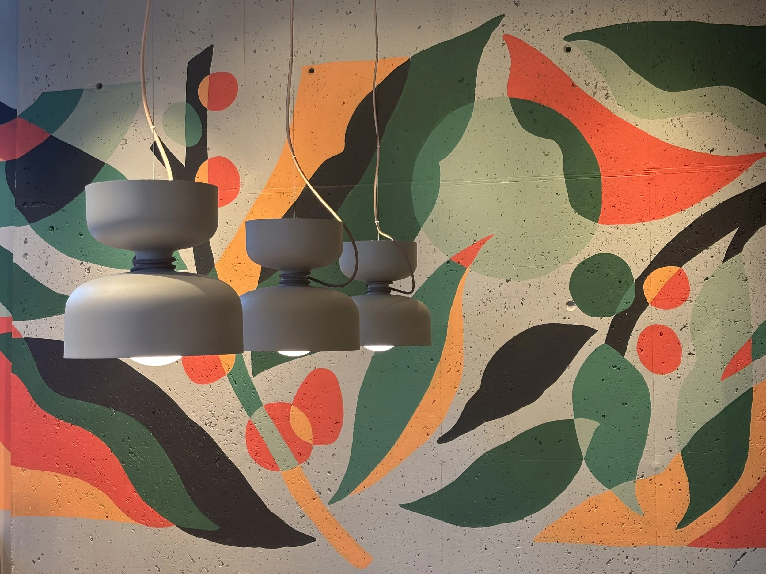 Colorful abstract mural with green, orange, black, and red shapes on a textured wall, with hanging white pendant lights in the foreground.