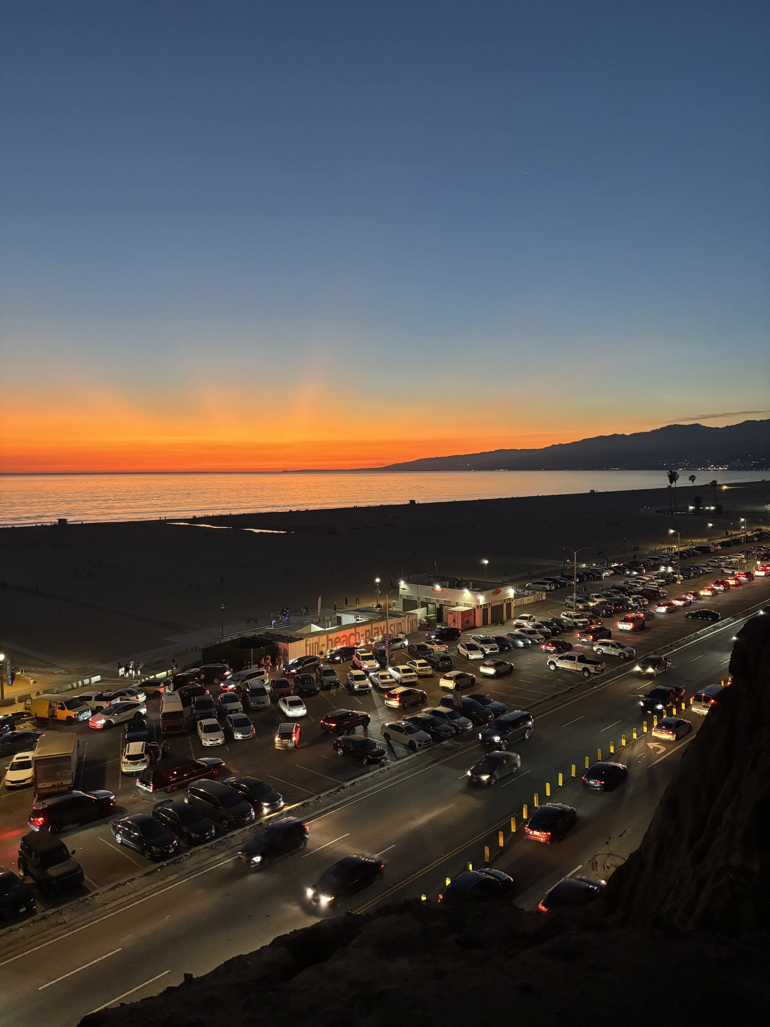 A beach parking lot filled with cars during sunset, with a view of the ocean and mountains in the distance.