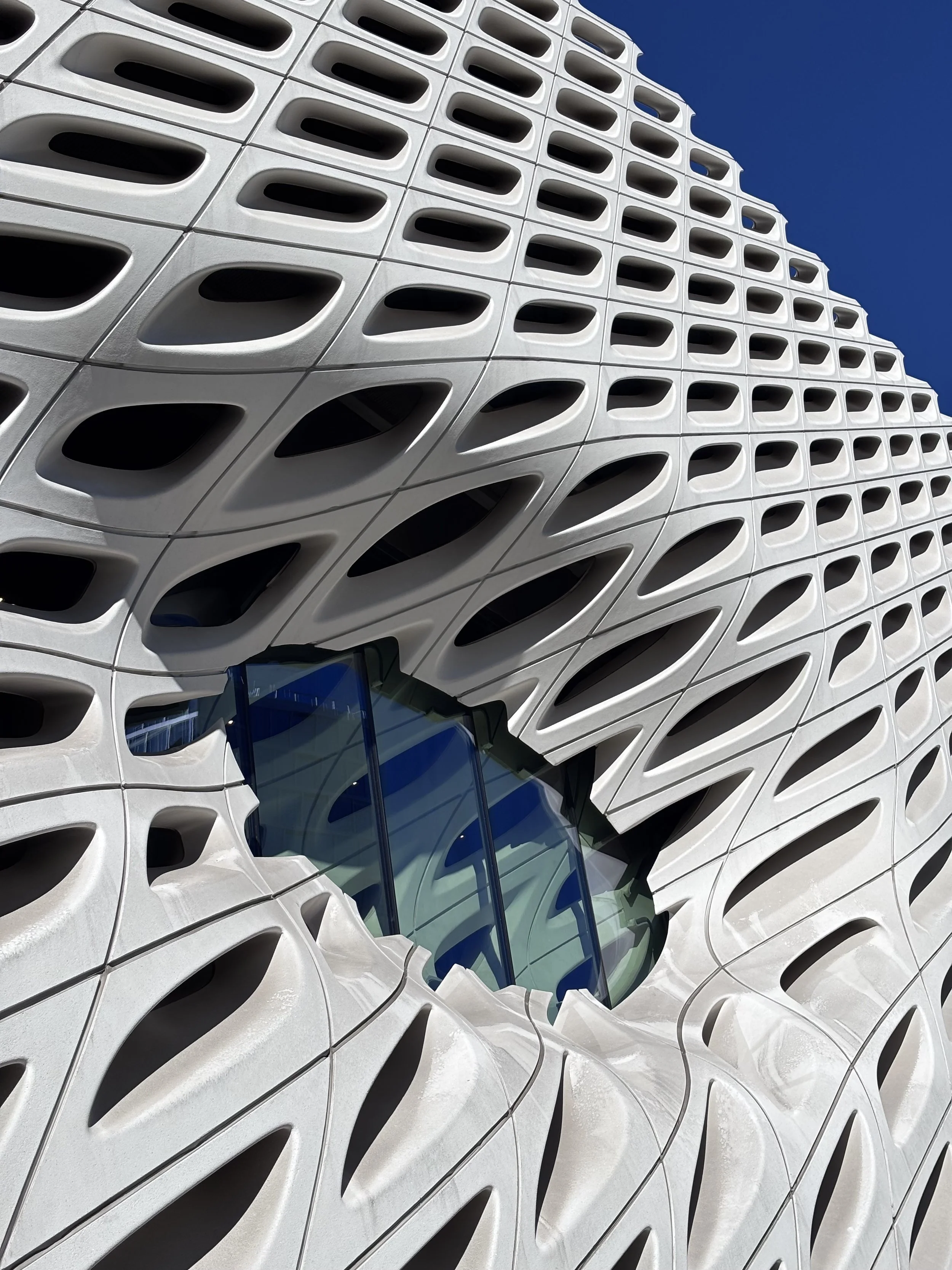 Close-up of a modern building's exterior with a geometric design of white, curved, lattice-like panels and reflective blue glass windows, under a clear blue sky.