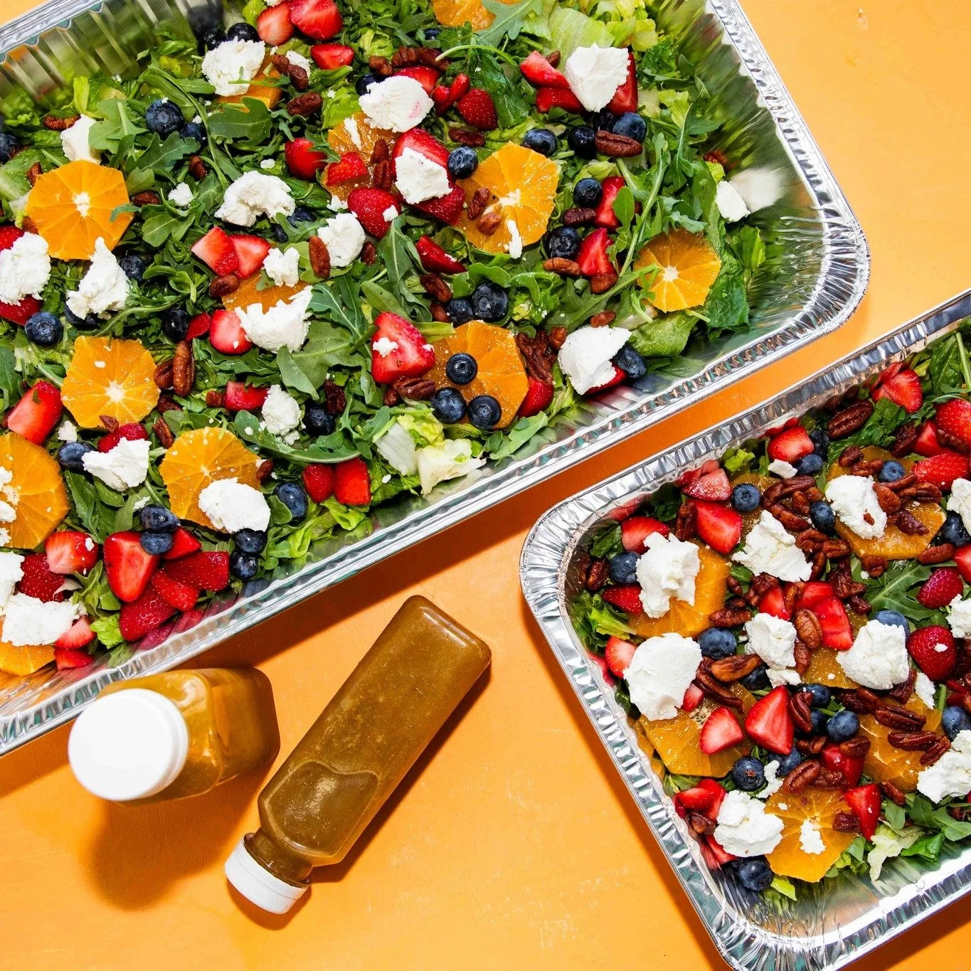 Two trays filled with fresh fruit salad containing strawberries, blueberries, orange slices, pomegranate seeds, and dollops of soft cheese, placed on an orange table. A bottle of honey or syrup and a squeeze bottle are also visible nearby.