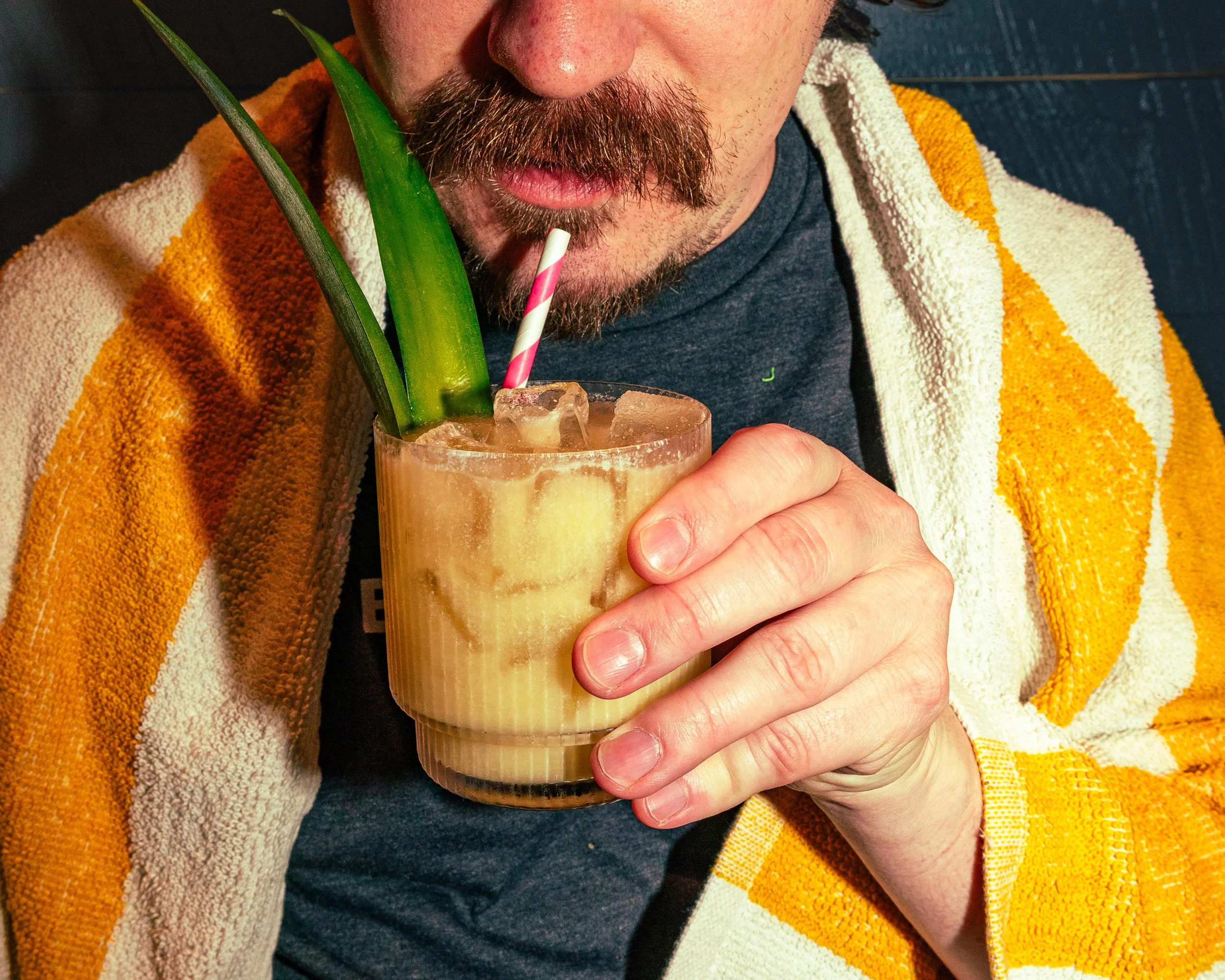 A man with a beard and mustache, wearing a dark shirt and a yellow and white striped towel over his shoulders, is holding a drink with a straw and a green leaf decoration, and is about to sip it.