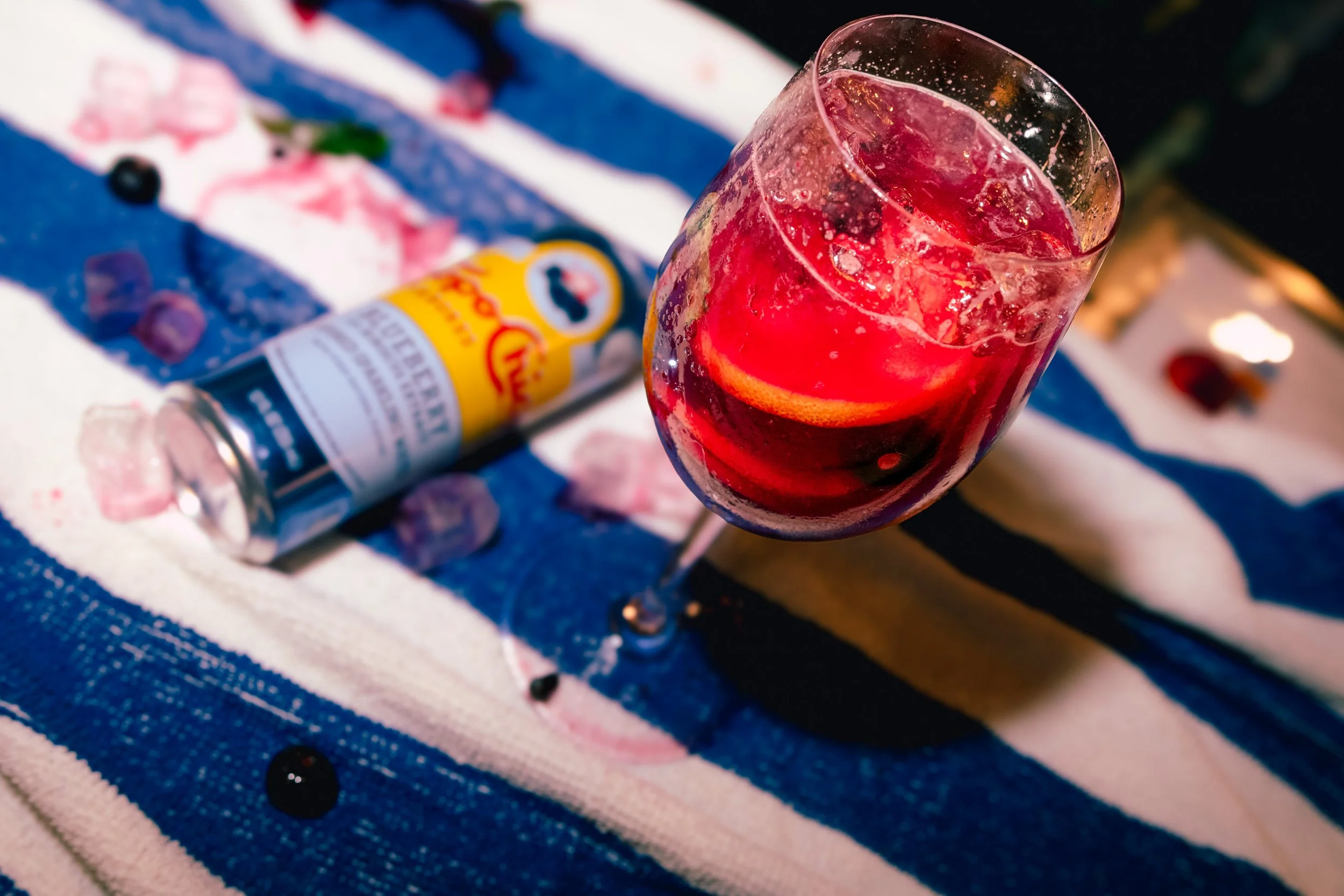 A glass of red soda with ice and a slice of orange on a striped blue, white, and pink cloth, with a bottle of flavored syrup in the background.