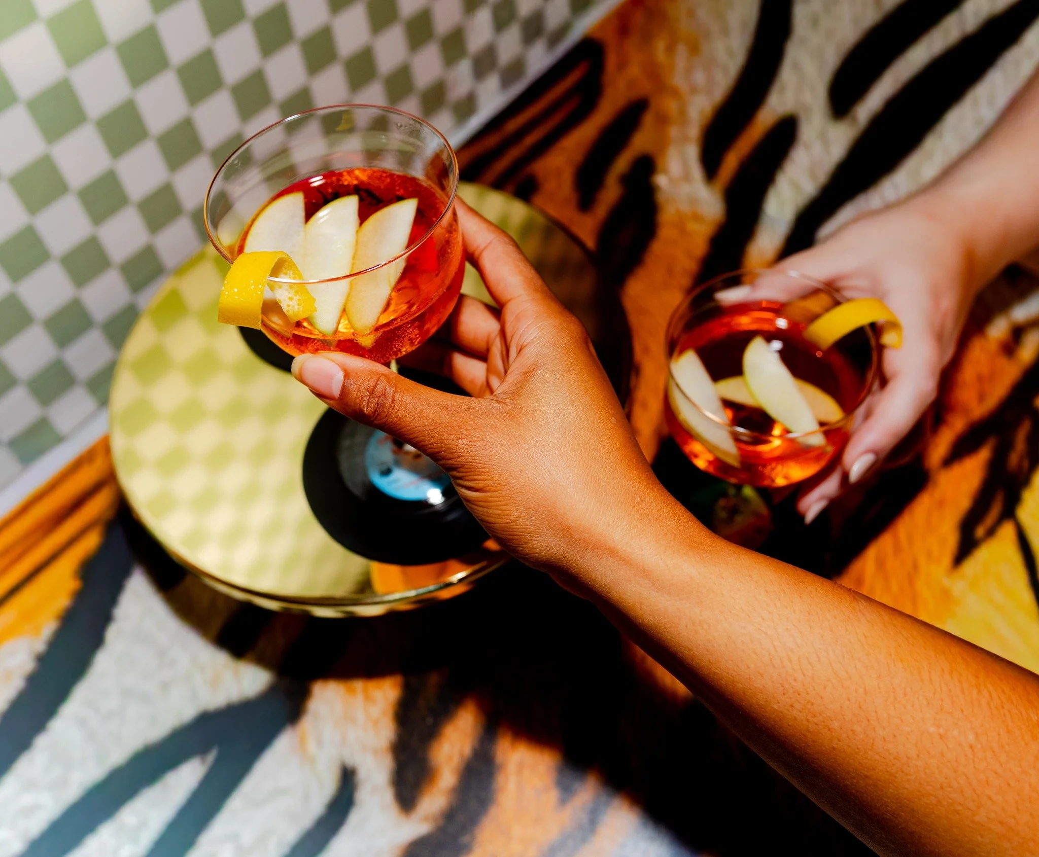 A person holding a clear glass of pinkish-red beverage with lemon slices and apple slices inside. The person is wearing a patterned shirt and has long-painted nails. The background features a checkered tablecloth with a bowl and a plate with food.