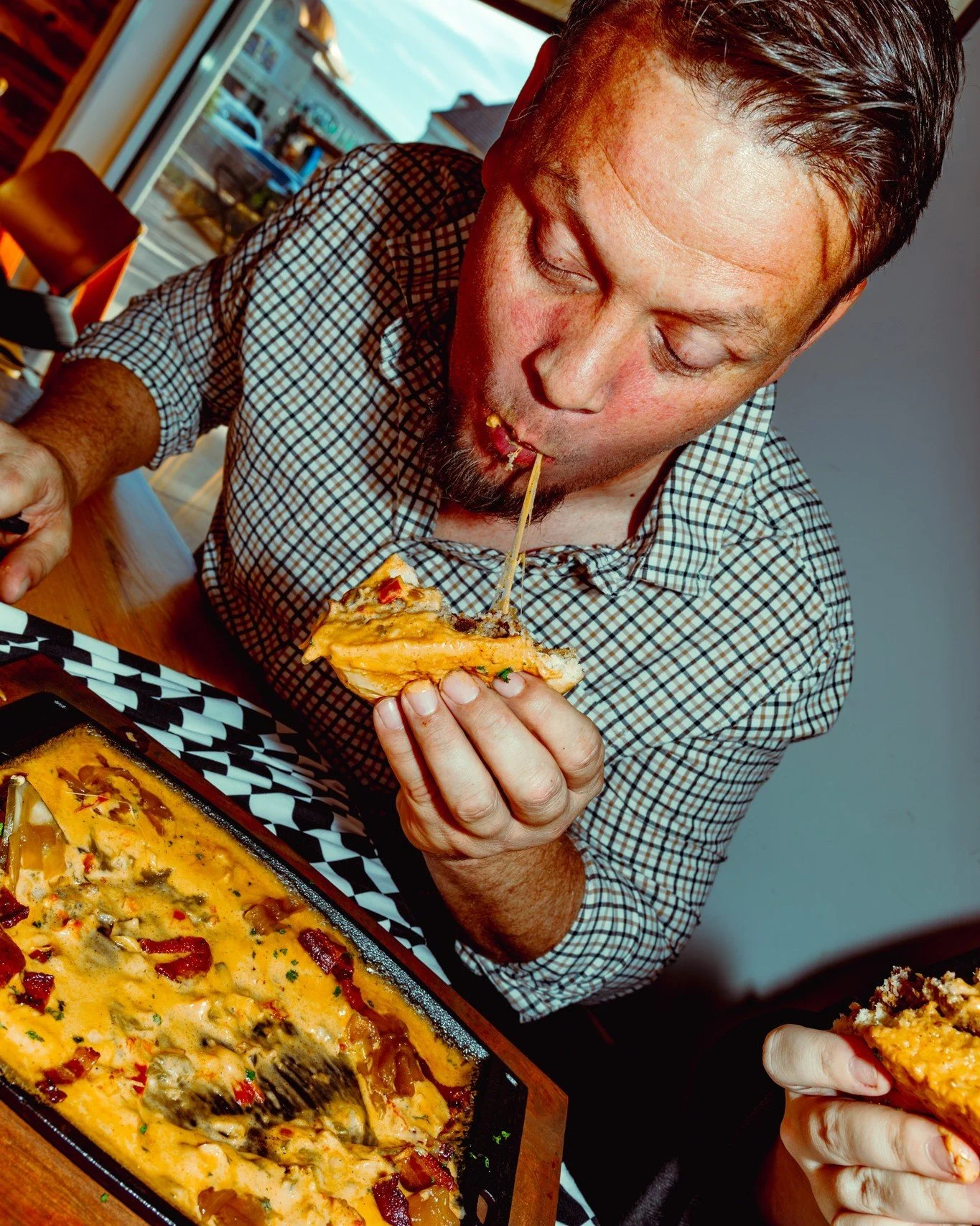 A man with short brown hair and a checkered shirt is eating pizza with cheese and toppings, sitting at a table with a burger dish in front of him.