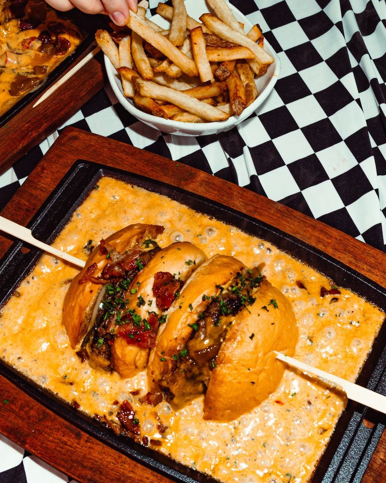 A burger topped with cheese and bacon on a skillet with a side of French fries in a bowl, served on a checkered tablecloth.