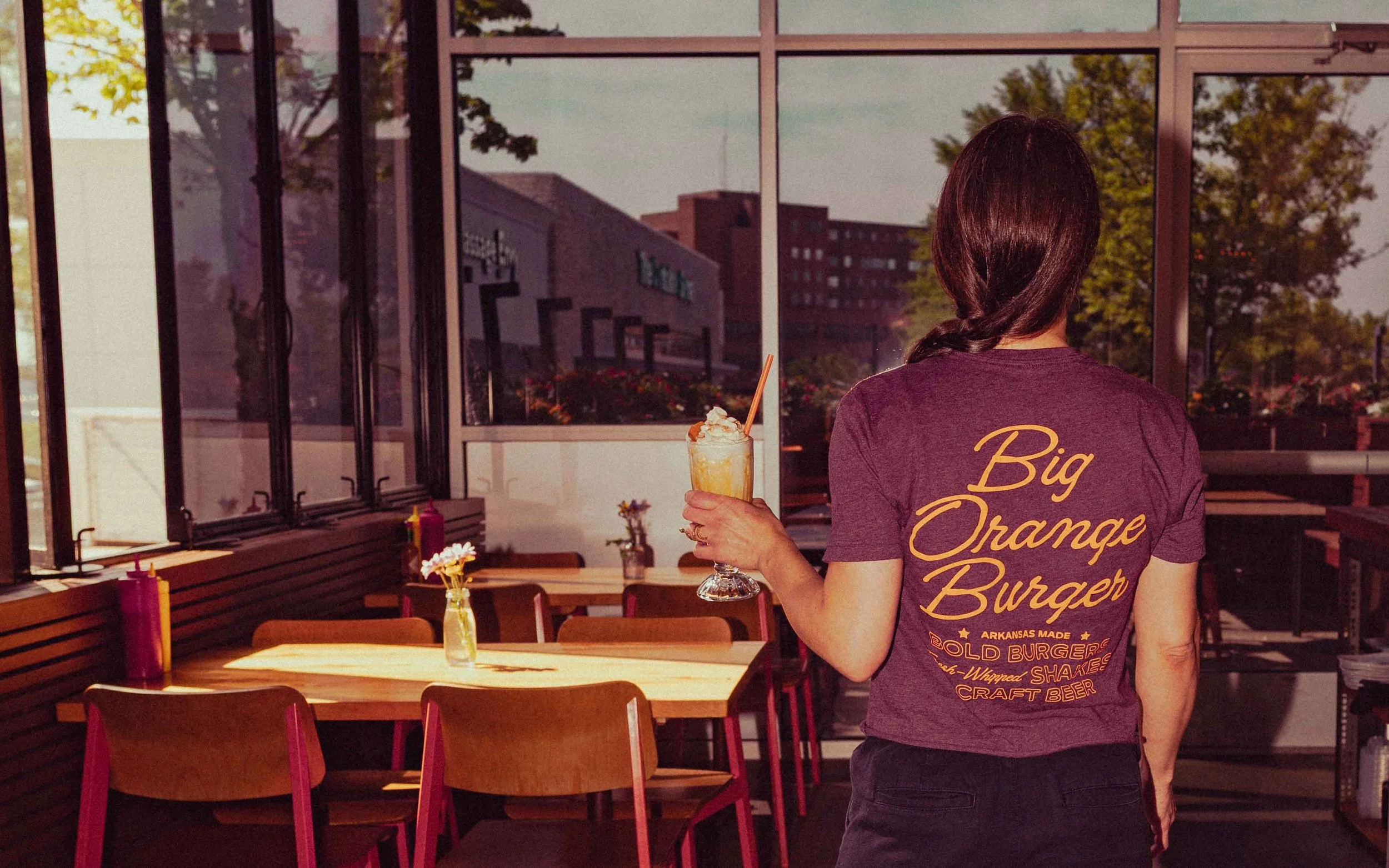 A woman with dark hair, seen from behind, wearing a maroon T-shirt that reads "Big Orange Burger" on the back, standing inside a restaurant holding a drink topped with whipped cream and a straw. The restaurant has large windows with a view of trees and buildings outside, with tables and chairs inside.