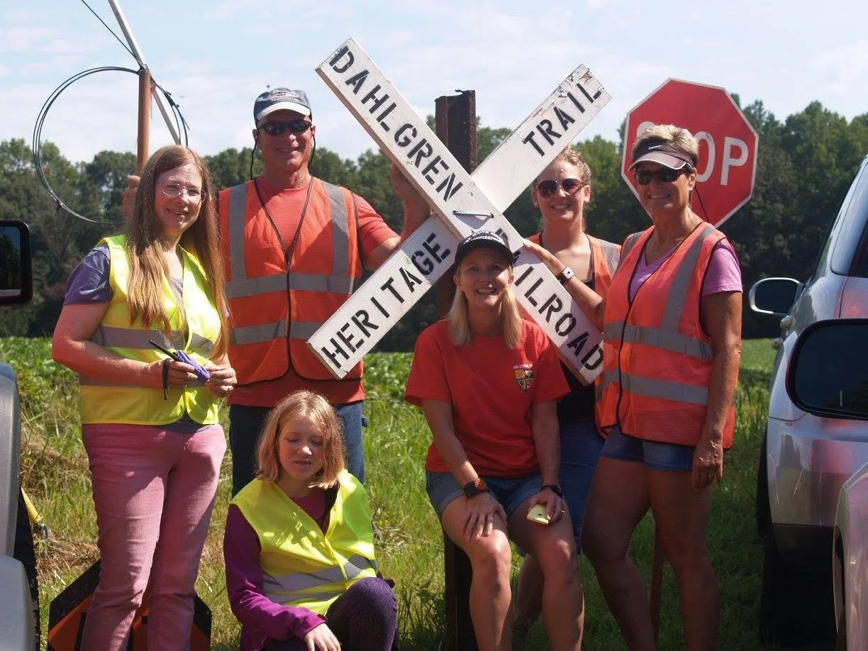 Group of seven people, five women and two girls, posing outdoors in front of a railroad crossing sign that says "Heritage - Dahle Green - Trail". Some are wearing safety vests, and a stop sign is visible in the background.