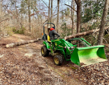 Person operating a green tractor with a front loader on a dirt trail in a wooded area.