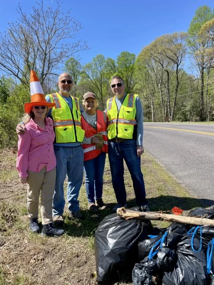 Four people standing on the side of a road, wearing safety vests, participating in a cleanup activity with trash bags and sticks, during a sunny day with trees in the background.