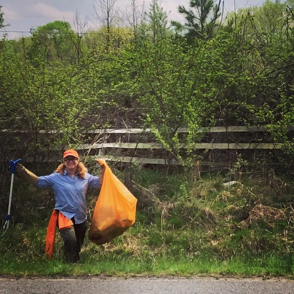 woman in outdoor clothes with trash bag and litter picker standing by roadside, smiling, with trees and an old wooden fence behind her.