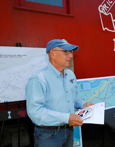 Man wearing glasses, a blue cap, and a light blue shirt holding a pamphlet, standing in front of maps on a red wall.