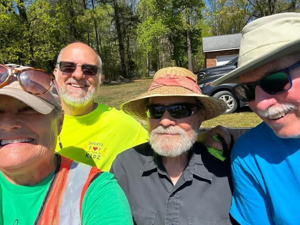 A woman and three men smiling and taking a selfie outdoors on a sunny day, with trees and parked cars in the background.