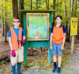 Two boys standing in front of an informational trail sign in a wooded area, both wearing orange safety vests, gloves, and casual clothing.