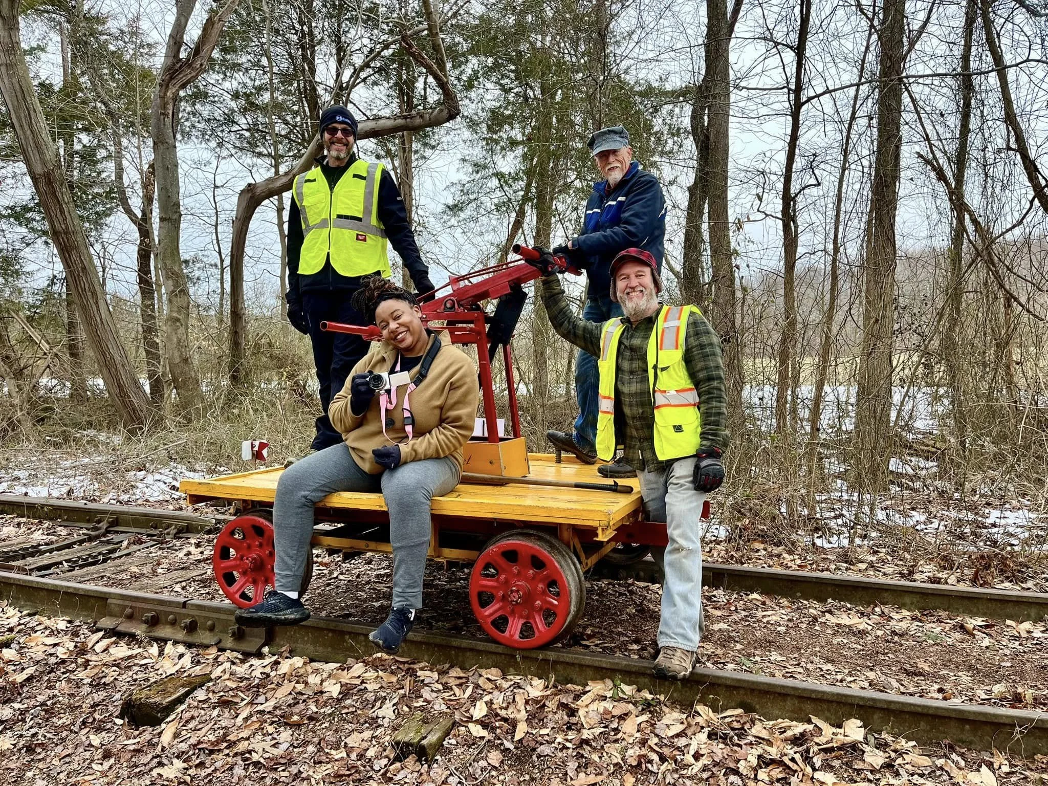 A group of five diverse people standing on a small train car on train tracks in a wooded area. They are smiling, some holding cameras, and appear to be enjoying an outdoor event or activity.