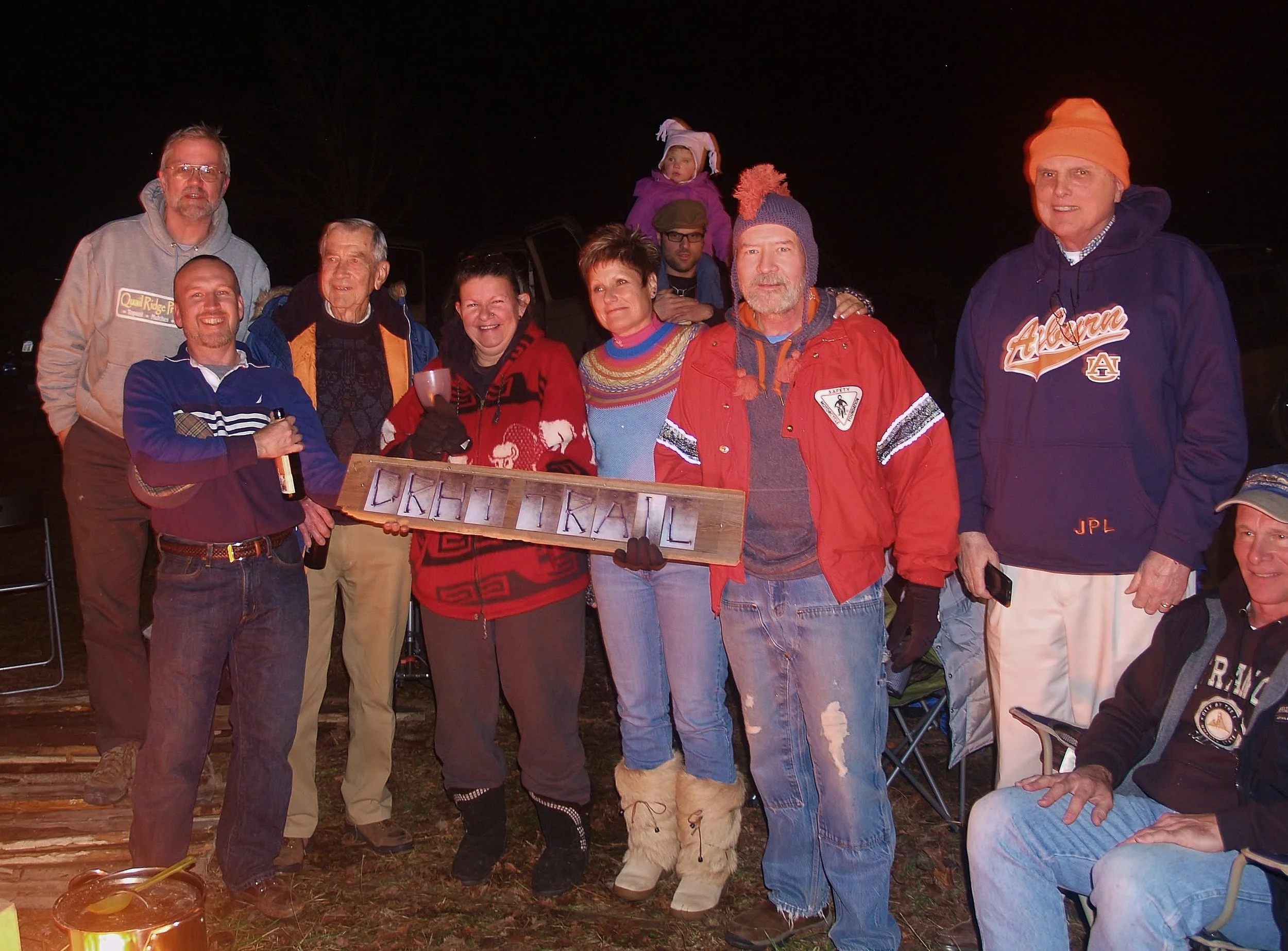 A group of people gathered outdoors at night, smiling and posing for a photo. Some are holding drinks, and there is a small bonfire on the ground in front. A person in the center is holding a wooden sign with the words 'Kikki Kral' on it.