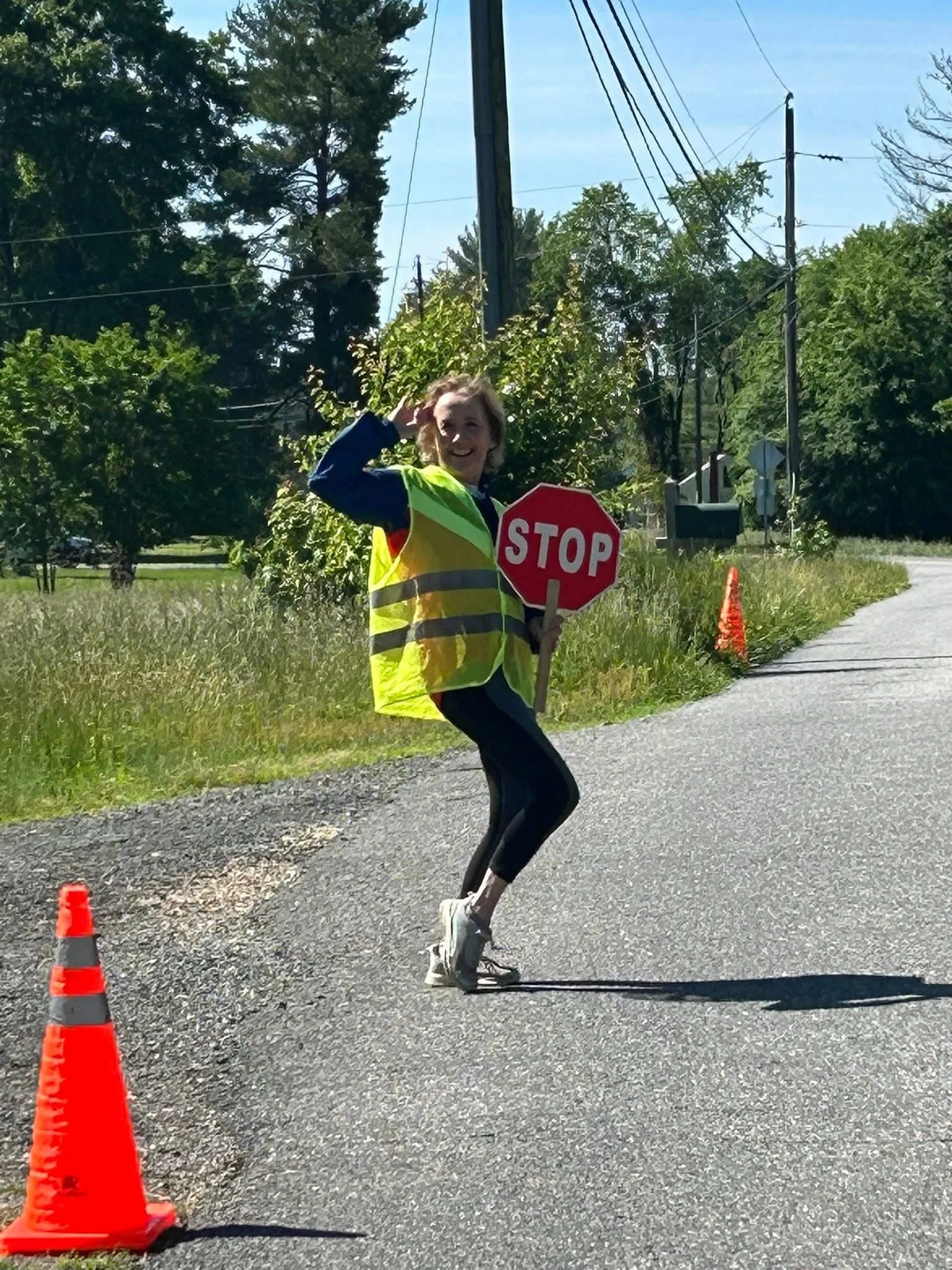 A person wearing a yellow reflective safety vest and black athletic clothing is running on a road, holding a red stop sign. They are smiling and waving with one hand. Orange traffic cones are placed on the side of the road, and trees with green foliage line the background.