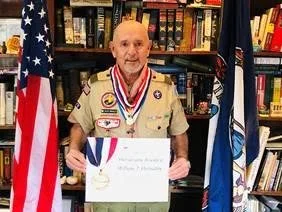 A man in a Boy Scouts uniform holding a certificate, standing in front of American and Boy Scouts flags with bookshelves in the background.