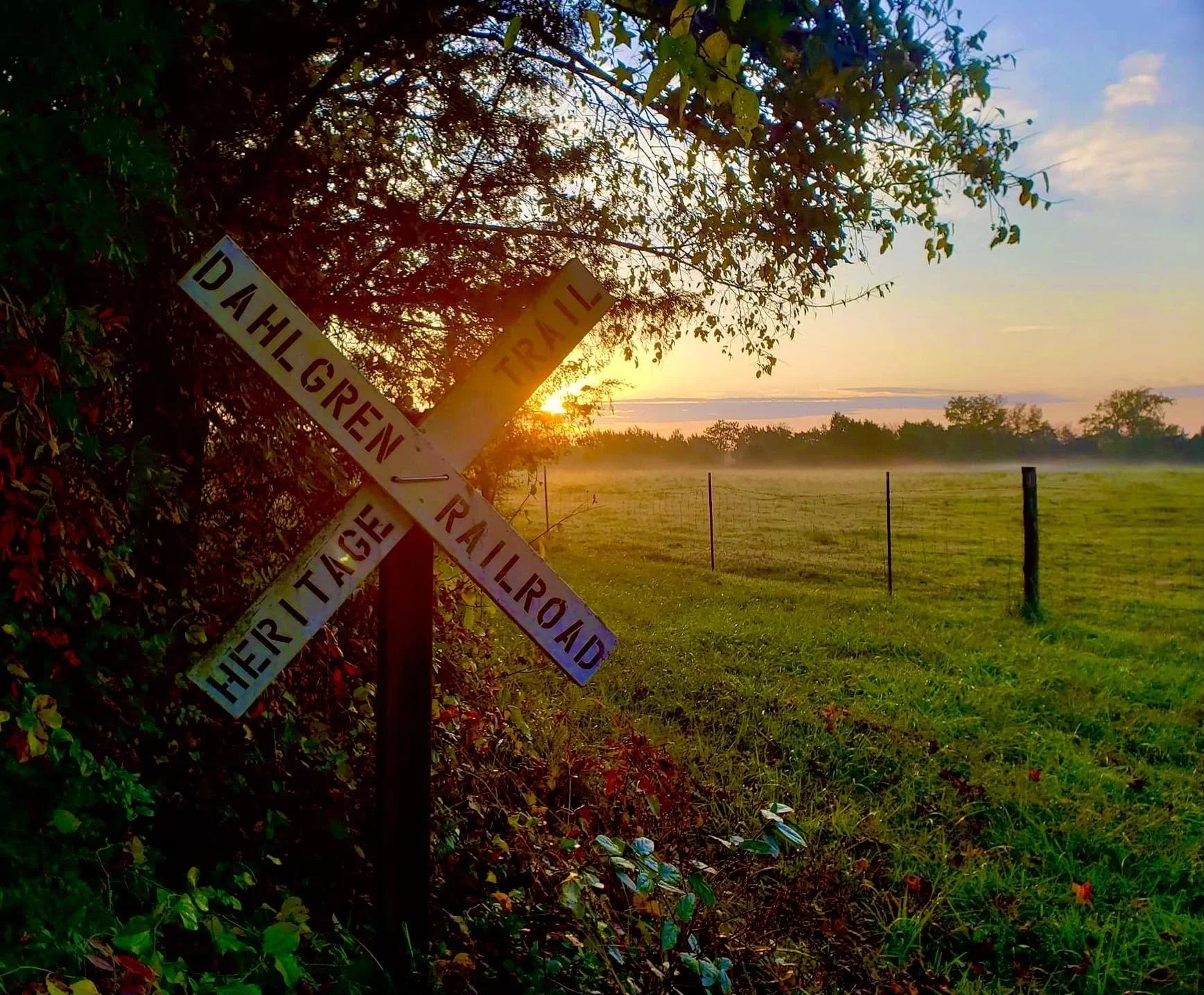 Sunset over a rural field with a barbed wire fence, through a hedge with a fallen railway crossing sign labeled 'Dahlegren Railway' in the foreground.